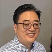 A man wearing glasses and a striped shirt, smiling, with a plain gray background.