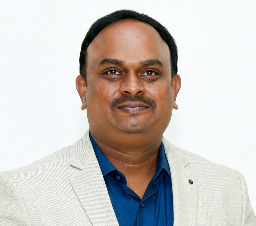 A man with medium skin tone, short black hair, and a thin mustache, dressed in a beige blazer and dark blue shirt, standing against a plain white background.