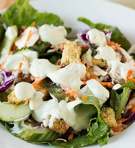A fresh garden salad with mixed greens, sliced cucumbers, shredded carrots, purple cabbage, croutons, and creamy dressing on top.