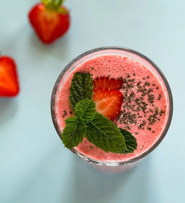 Pink smoothie garnished with sliced strawberry, fresh mint leaves, and chia seeds, with whole strawberries in the background on a light blue surface.