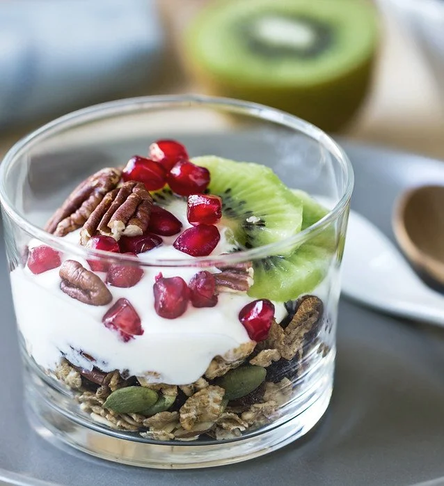 A glass of yogurt parfait topped with pomegranate seeds, kiwi slices, pecans, and granola with a blurred kiwi in the background.