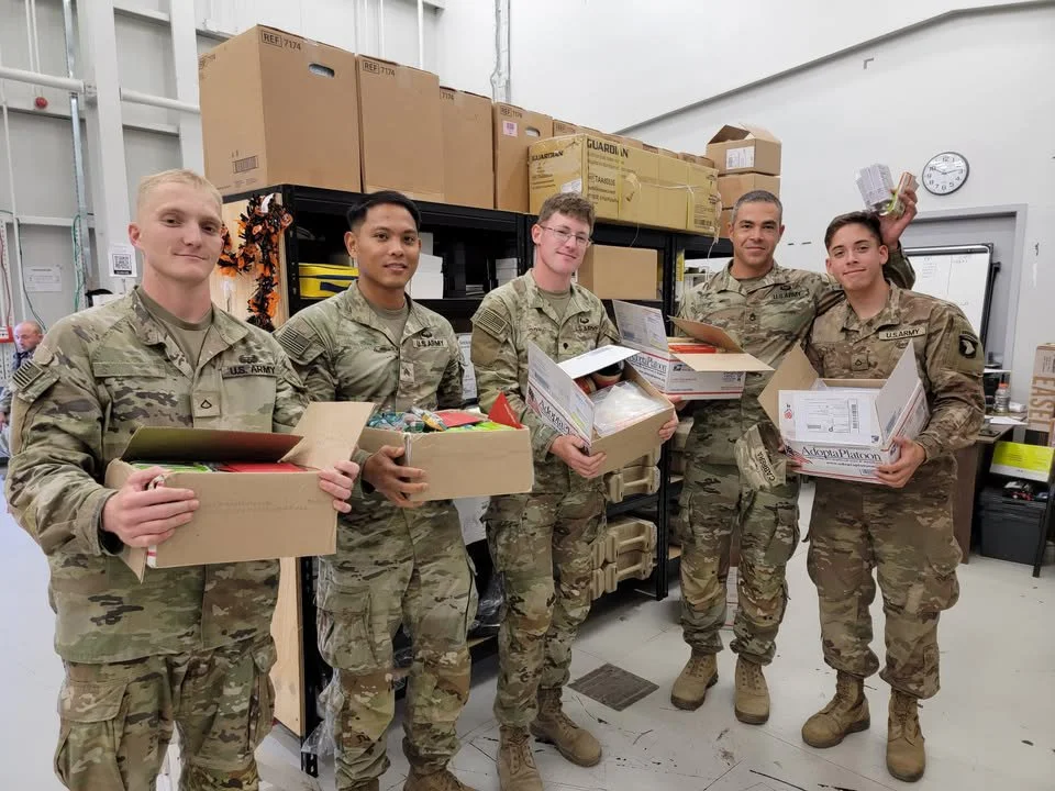 Five U.S. Army soldiers standing in a room holding boxes filled with donations or supplies.