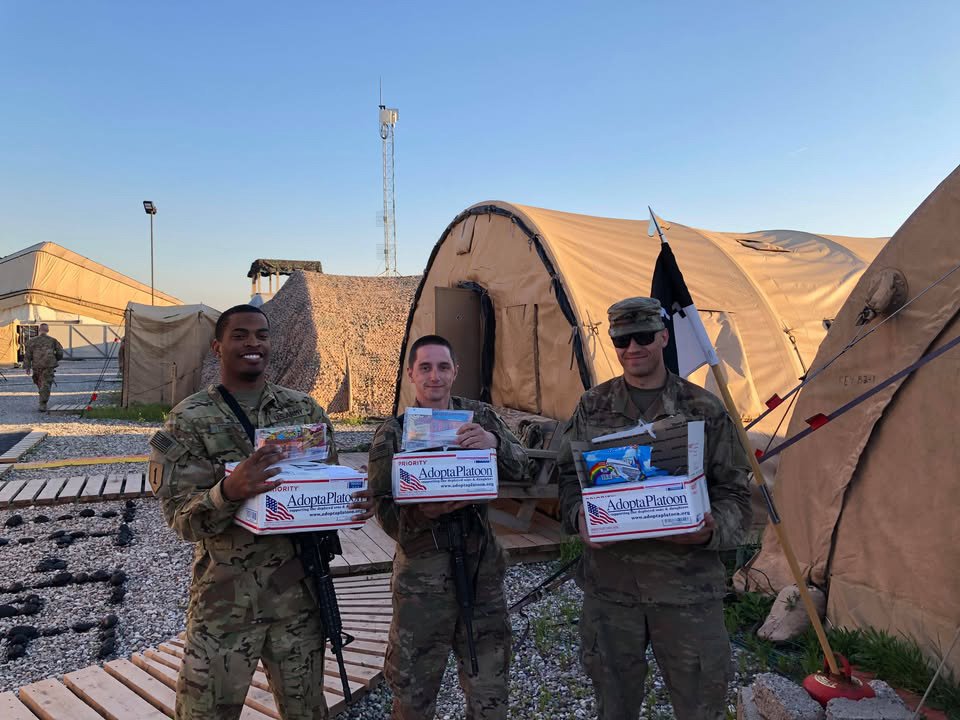 Three soldiers in camouflage uniforms standing in front of tents, each holding a box labeled 'Adopt a Platoon,' with one soldier smiling widely and the other two wearing sunglasses, in a military base setting.