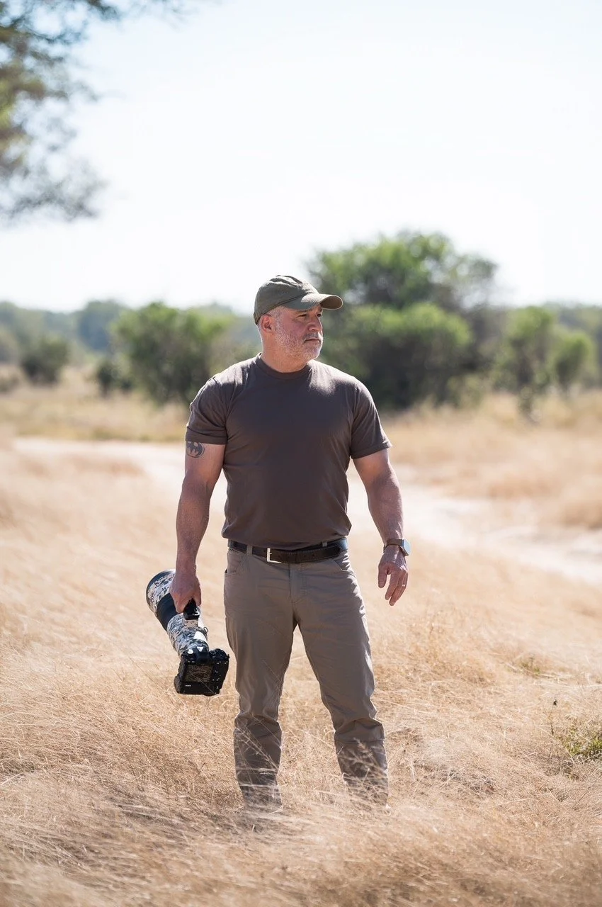 A man walking outdoors in a dry, grassy landscape with trees in the background, holding a large camera lens in his right hand, wearing a gray t-shirt, khaki pants, a cap, and a watch.