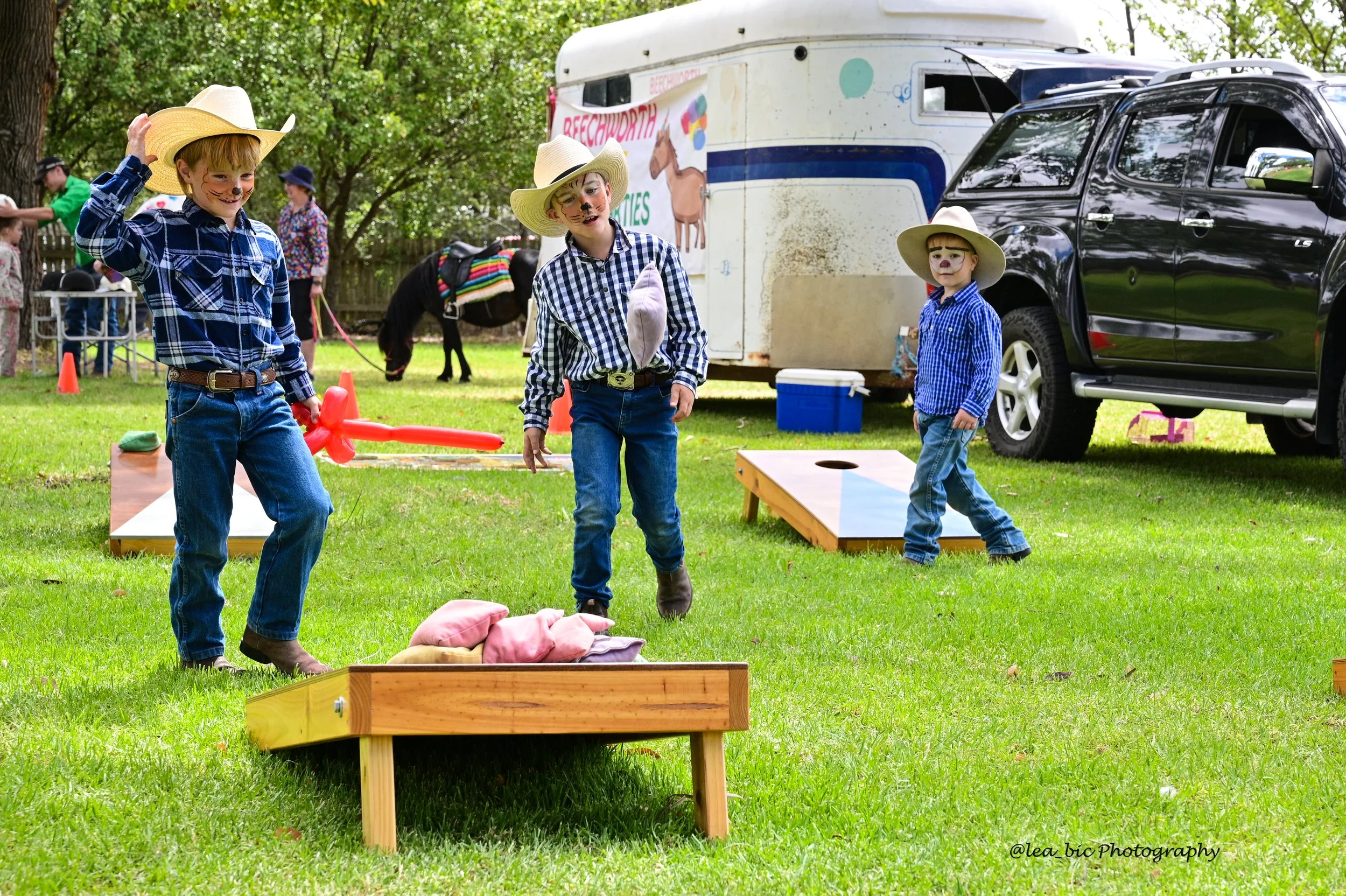 Three children dressed as cowboys playing a cornhole game at Holbrook Races, outdoors with a green background, trailer, and car in the distance.
