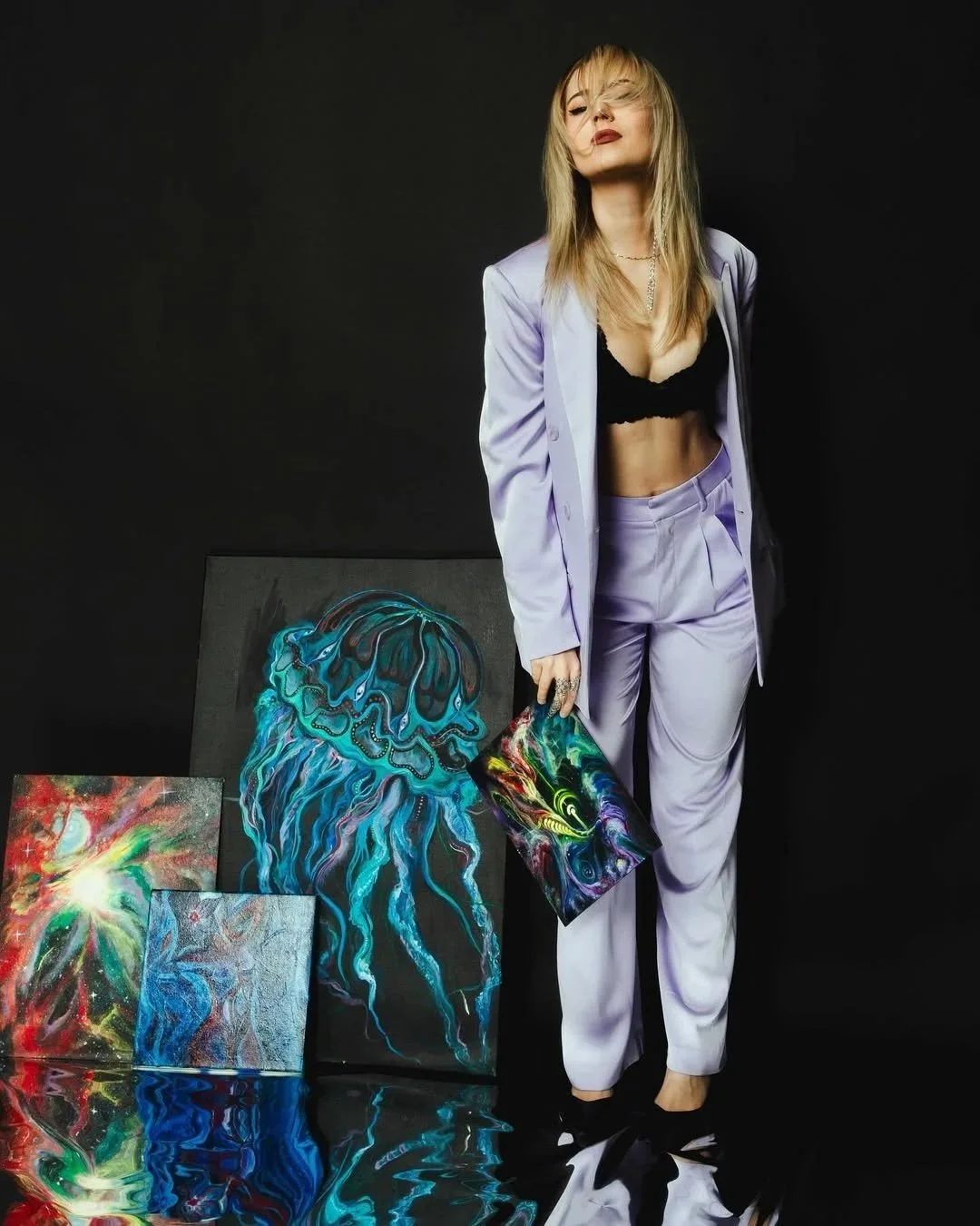 A woman with blonde hair wearing a lavender suit, black bra, and black shoes standing in front of black background, holding a colorful abstract art piece, with additional paintings on the floor, including a jellyfish painting.