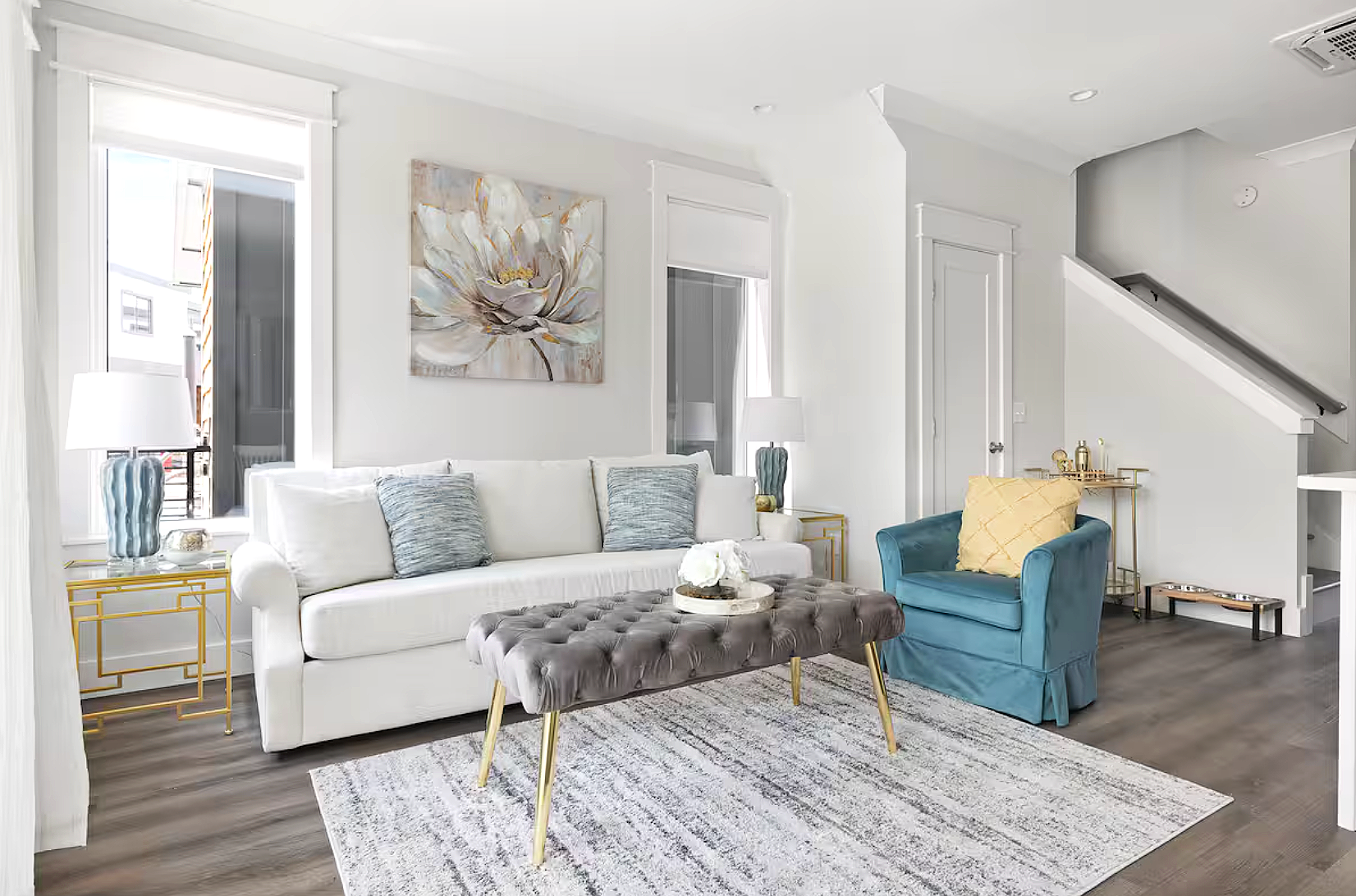 Living room with white sofa, blue armchair, gray tufted ottoman, and gold side tables with lamps. Wall art of a flower hangs above the sofa. Window with white blinds and a sliding door to an outdoor balcony. Light-colored area rug on dark wood flooring.