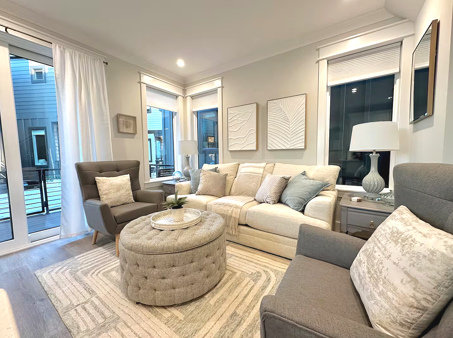 Living room with beige sofa, gray armchair, ottoman, side tables, lamps, artwork, and large windows with white curtains.