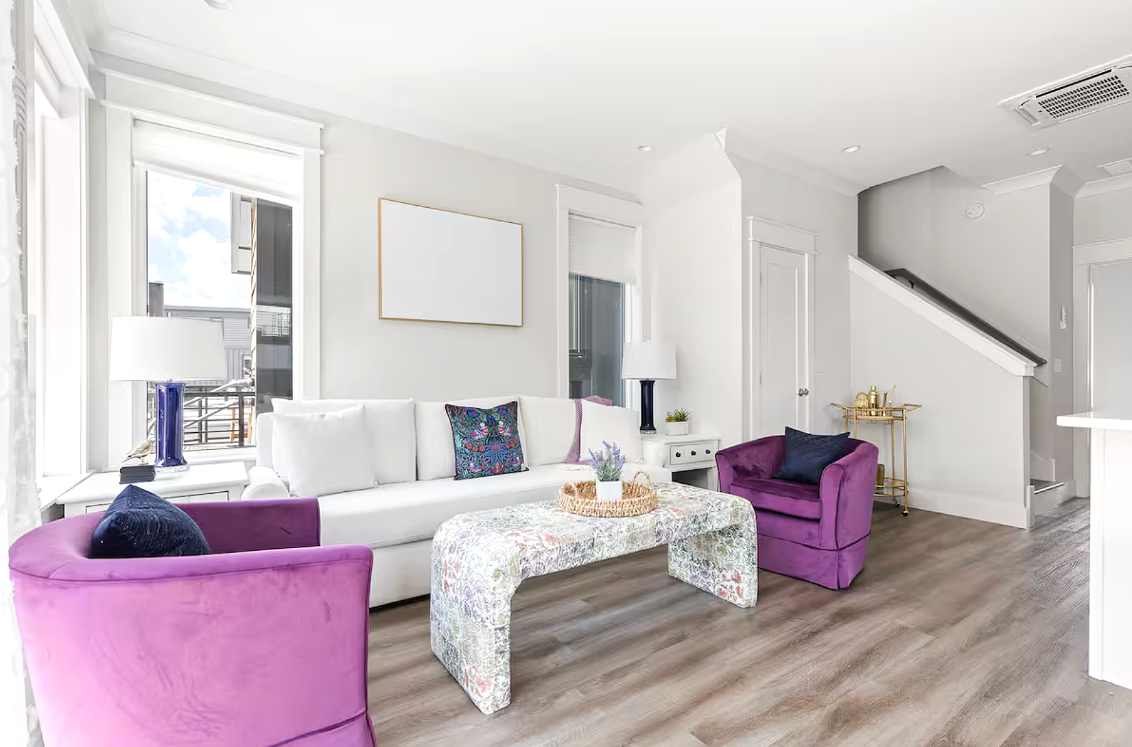 Bright living room with white couch, two purple armchairs, a floral-patterned coffee table, and decorative pillows. Large windows with white trim provide natural light. Modern decor includes lamps, small plants, and artwork. Hardwood floors and white walls create a clean, airy atmosphere.