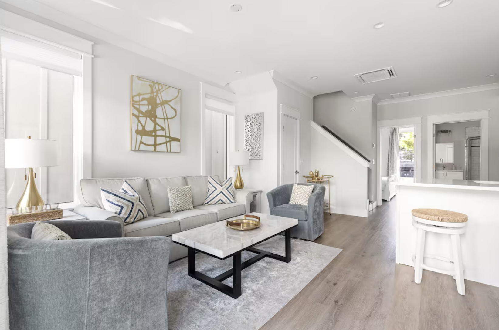 Bright living room with a white sofa, gray armchairs, abstract wall art, white and gold lamps, a marble coffee table, and large windows with blinds.
