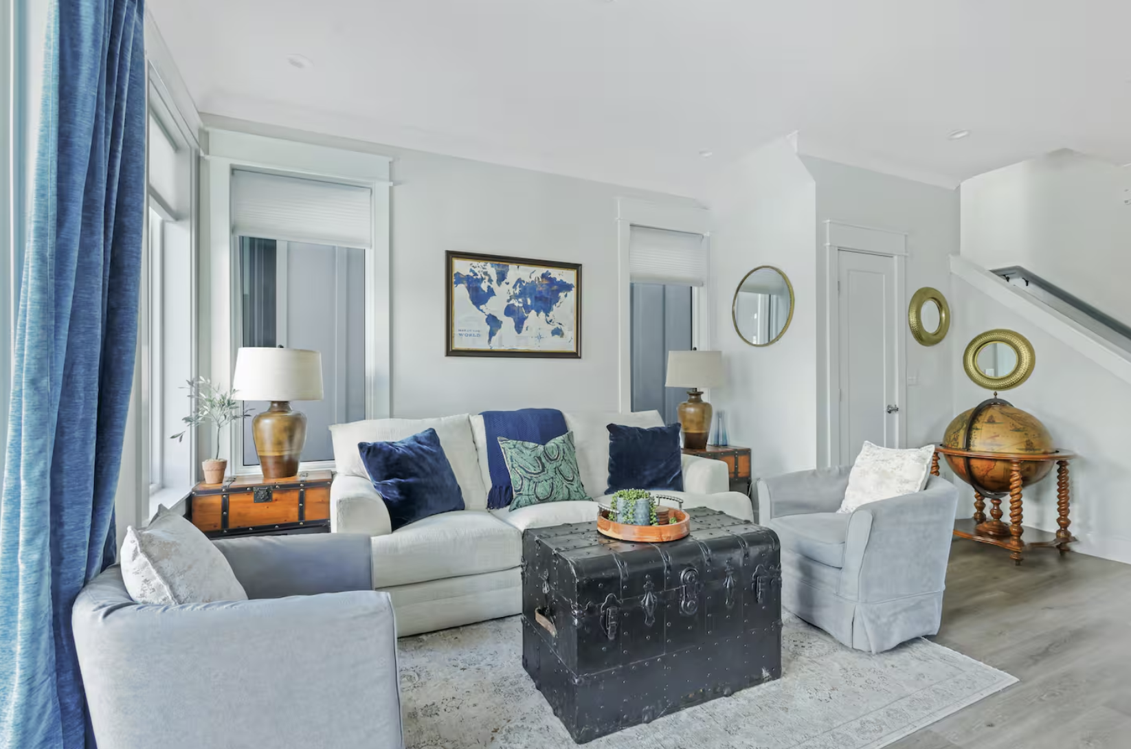 Living room with light-colored sofa, armchairs, wooden side tables, blue curtains, world map art, round mirrors, globe, and decorative pillows.