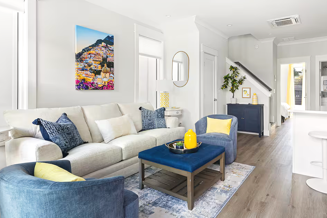 Bright living room with white sofa, blue armchairs with yellow pillows, a dark blue coffee table, and colorful wall art. Bright windows, a staircase, and decorative accents are visible.