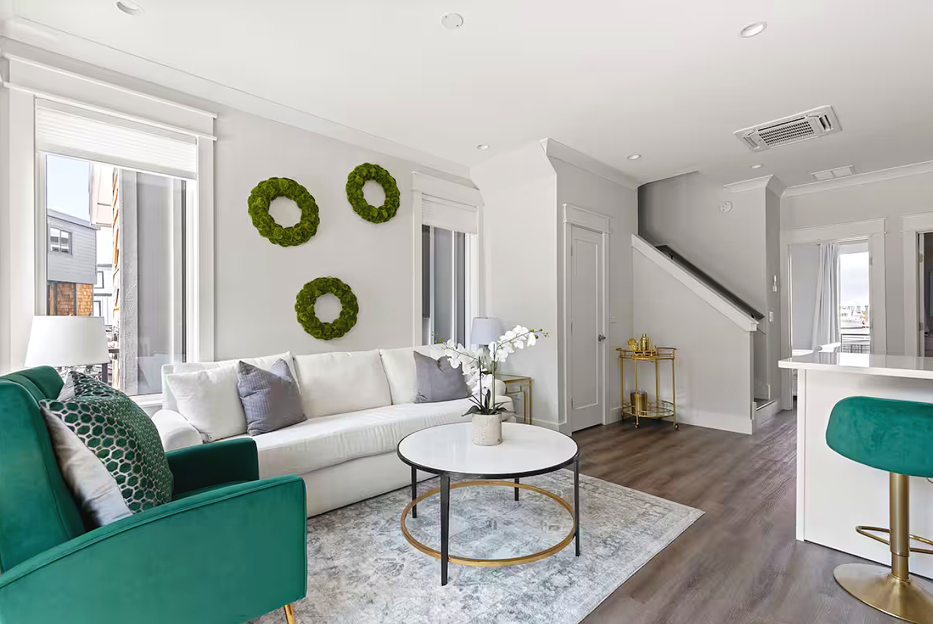 Living room with white sofa, green armchair, round marble coffee table, flower arrangement, white walls with three green wreaths, windows with white blinds, hardwood floors, staircase, and partial view of kitchen.