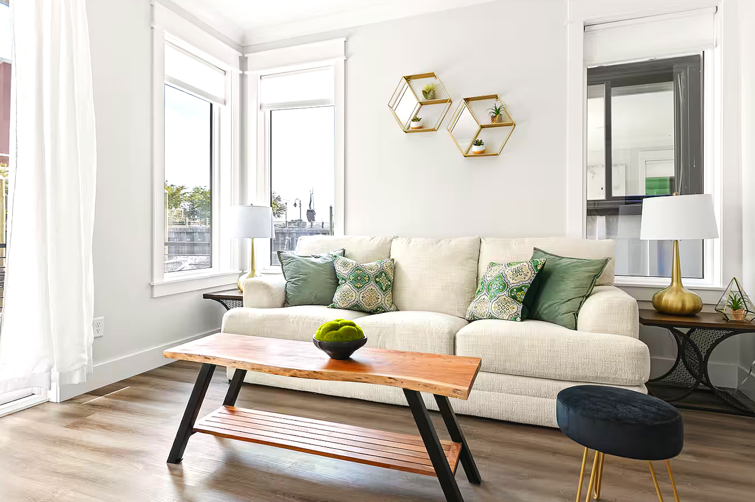 Bright living room with large windows, white walls, beige sofa with green and patterned cushions, wooden coffee table with a bowl of green moss, side tables with gold lamps and decor, wooden floor.