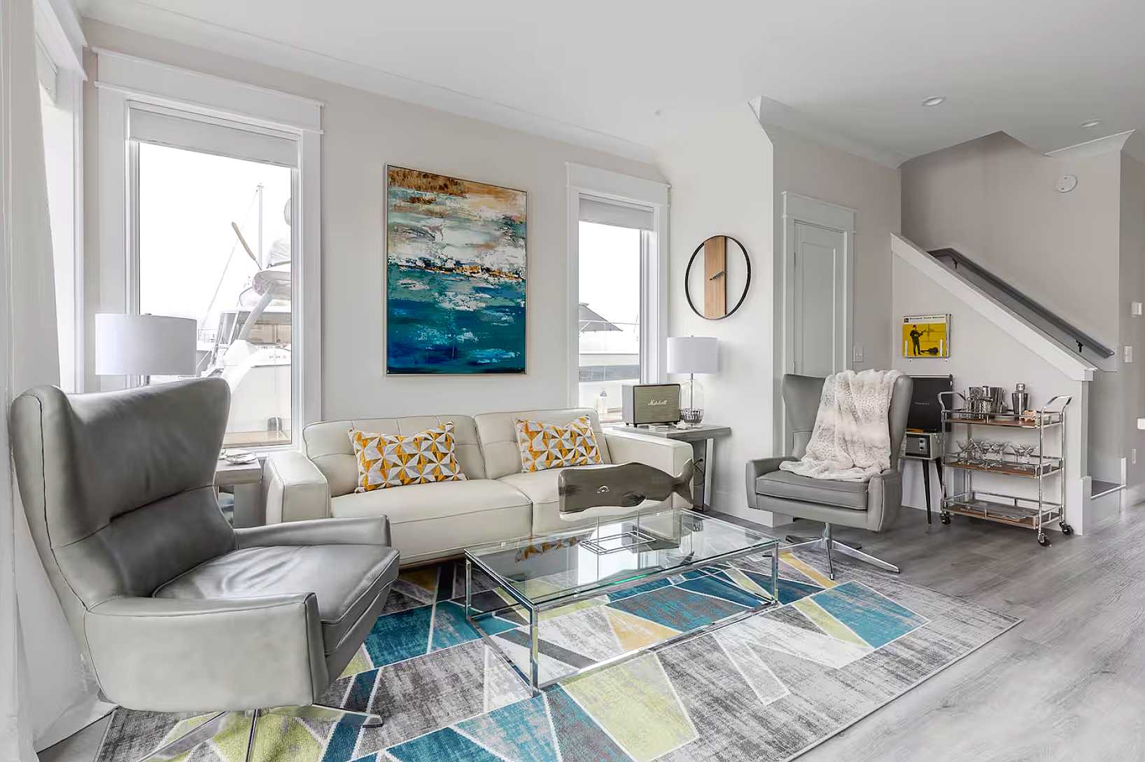 Modern living room with white sofa, gray armchair, glass coffee table, colorful geometric area rug, paintings, and large windows.