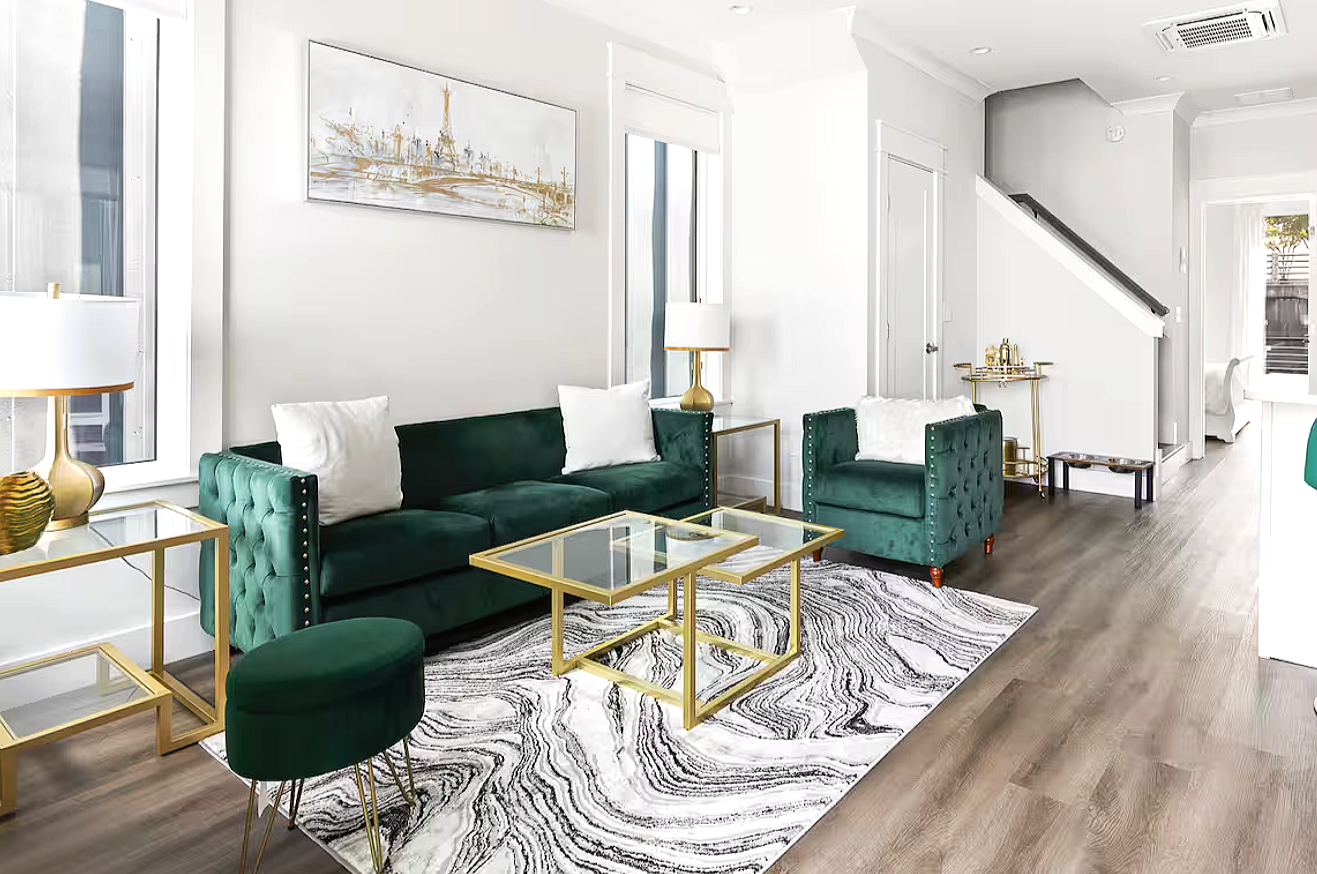 Bright living room with white walls, green velvet sofa and armchair, gold-framed glass coffee tables, black and white marbled rug, white curtains, and various decorative items.