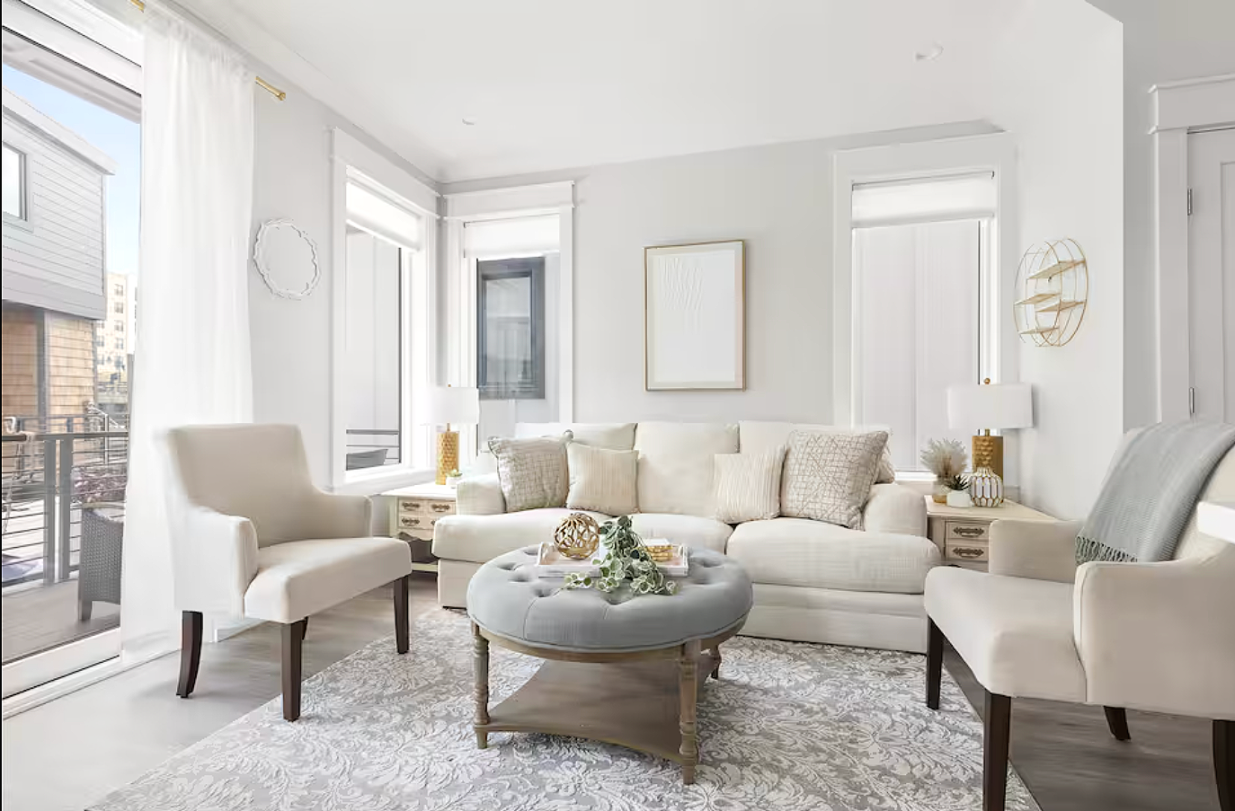 Bright living room with white walls, beige furniture, and large windows letting in natural light, decorated with neutral-toned pillows, a rug, and minimalist wall art.