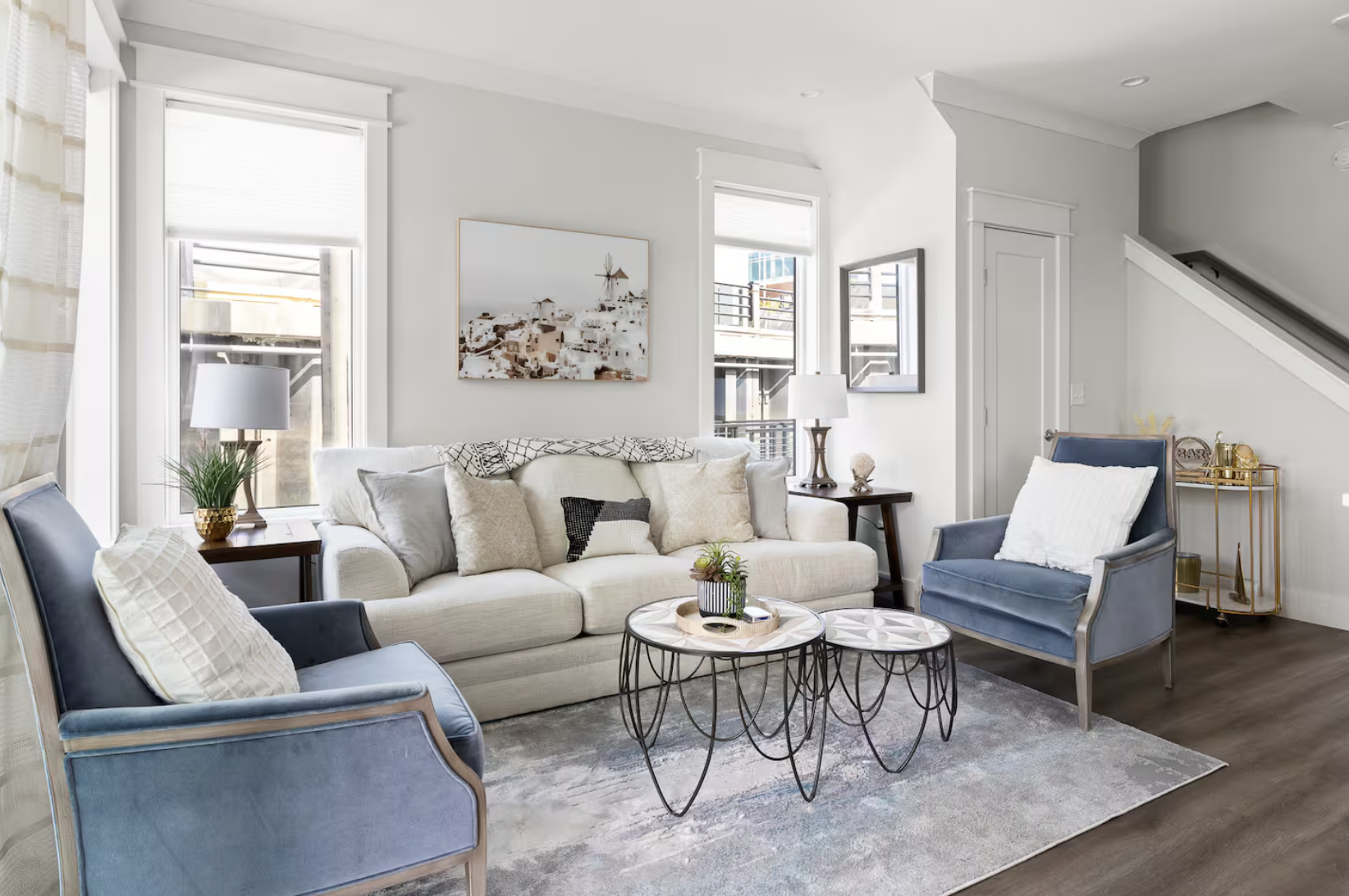 Living room with white sofa, blue armchairs, light wood side tables, and modern decor with windows and wall art.