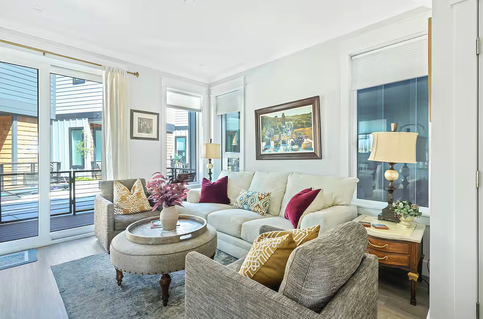 Living room with white sectional sofa, beige and gray armchairs, colorful pillows, a round ottoman with a tray and vase with pink flowers, windows with sheer curtains, a wall painting, and a side table with a lamp and plant.
