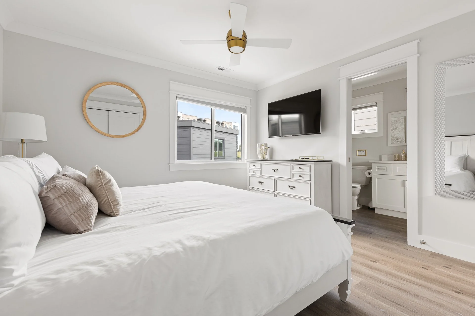 A bedroom in wilmington nc with a white bed, beige pillows, a white dresser, a wall-mounted TV, a window view of neighboring buildings, and an adjacent bathroom with white cabinetry.