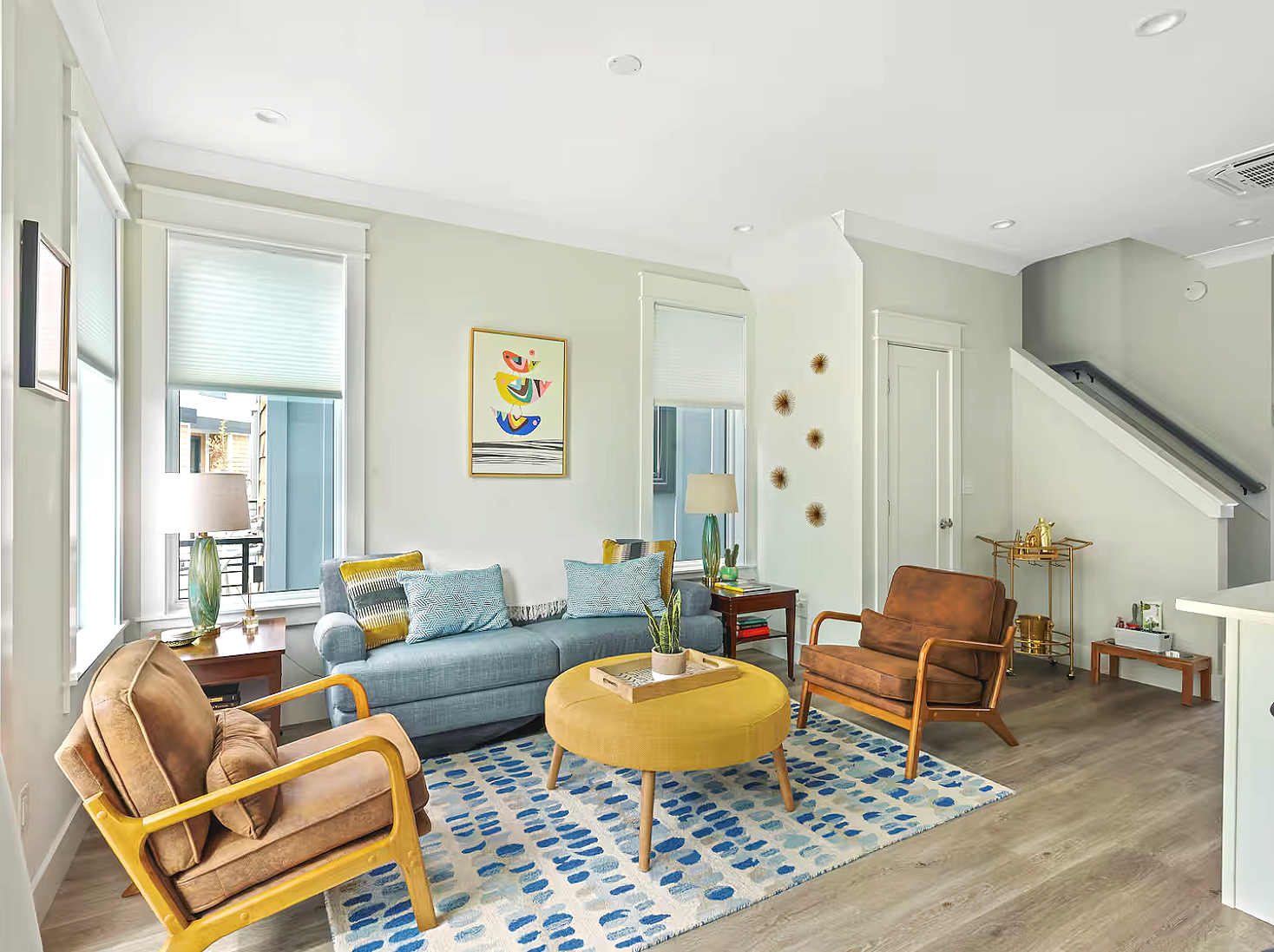 Bright living room with large windows, a light blue sofa, colorful pillows, a yellow ottoman, a brown armchair, a modern wooden chair, and blue patterned rug; decorated with wall art, lamps, and houseplants.