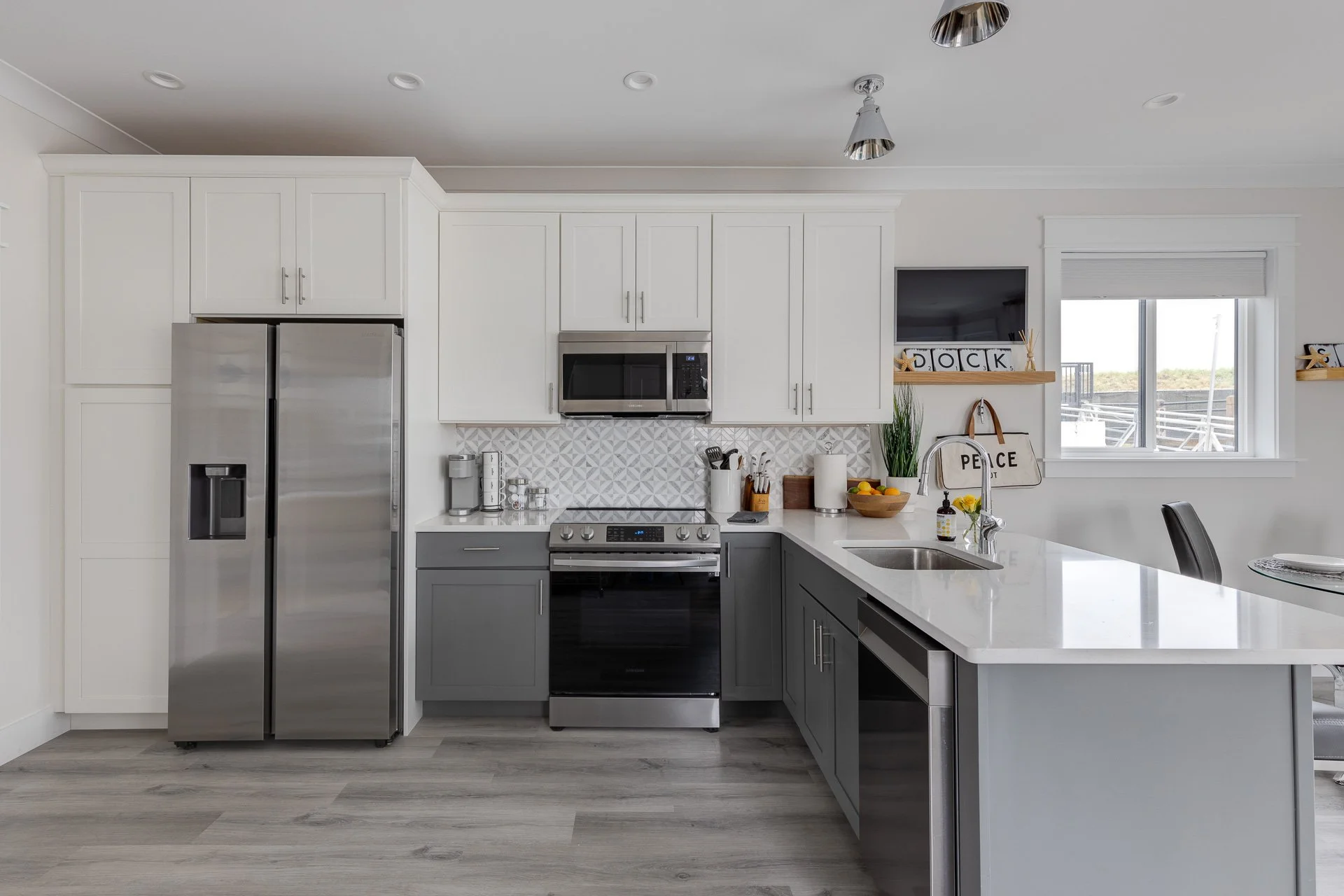 Modern kitchen with white upper cabinets, gray lower cabinets, stainless steel refrigerator, microwave, oven, and a white countertop with a sink, window, and decorative items.