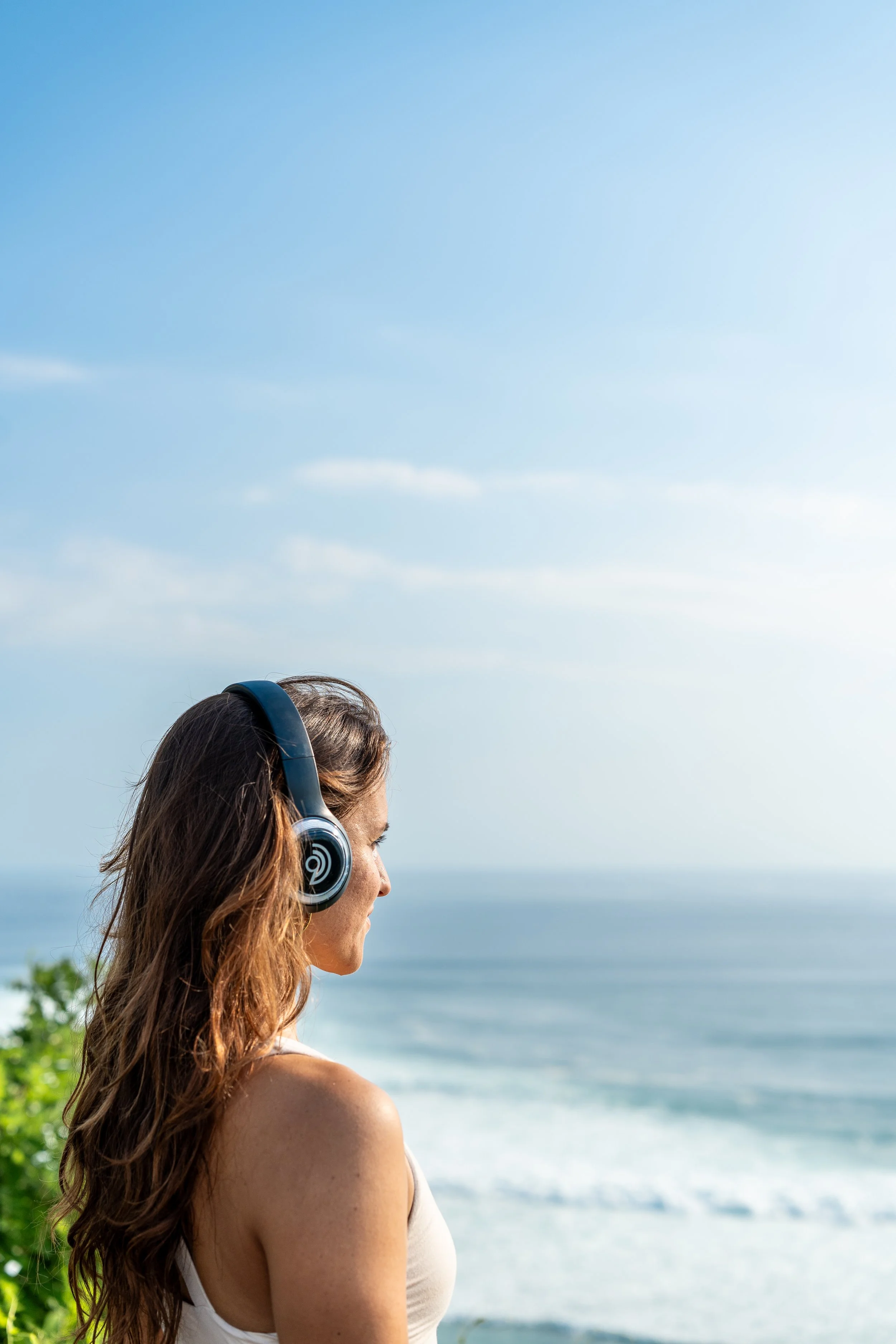 A woman with long brown hair wearing headphones, standing outdoors near the coast with ocean waves and a blue sky in the background. Holotropic Breathwork