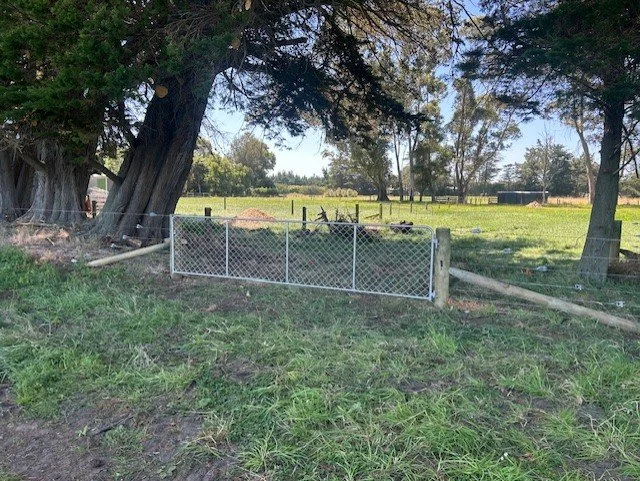 A fenced area in a grassy field with trees and open space, featuring a large tree leaning over the fence and some smaller trees and structures in the background.