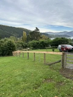 A grassy field with a wooden fence, trees, mountains in the background, and cloudy sky.