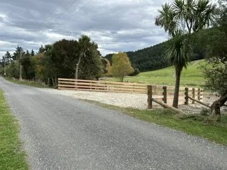 A rural road with a wooden fence, green trees, and hills under a cloudy sky.