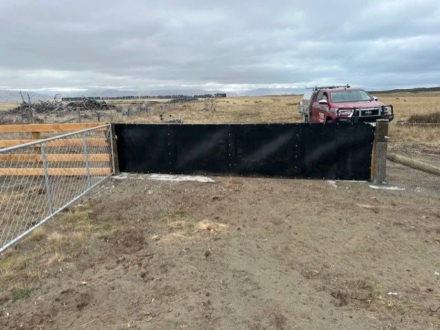 A black outdoor gate with a red pickup truck parked behind it, in a rural open field with cloudy skies.