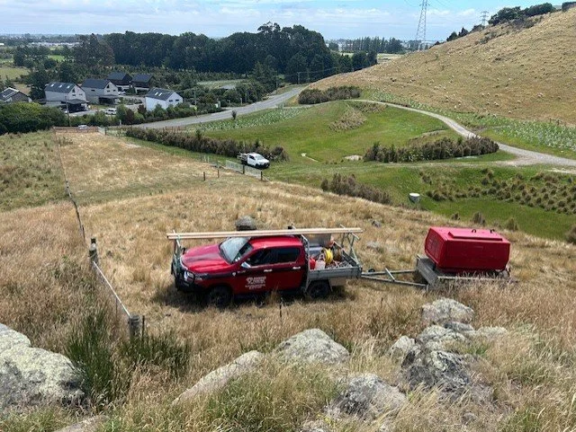 A red utility truck with a ladder on top is parked on a grassy hillside, towing a red storage container on a small trailer.