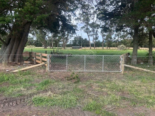 A chain-link gate with wooden fencing on either side, set in a rural outdoor area with trees and green fields in the background.
