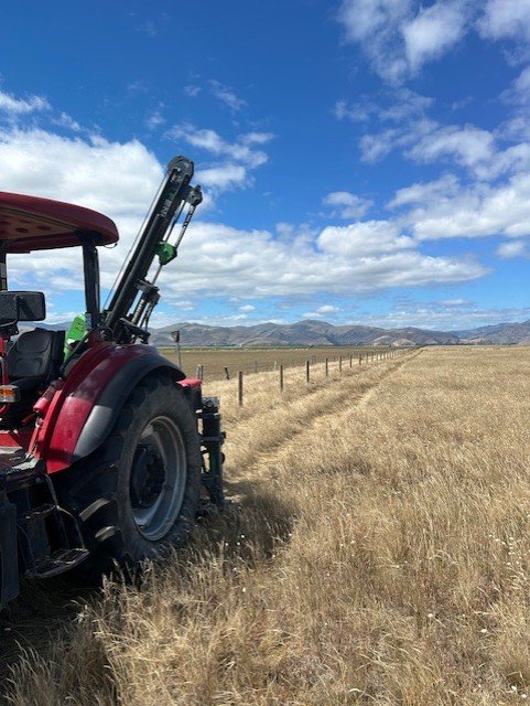 A red tractor with a green attachment on a grassy field under a blue sky with clouds, distant mountains in the background.