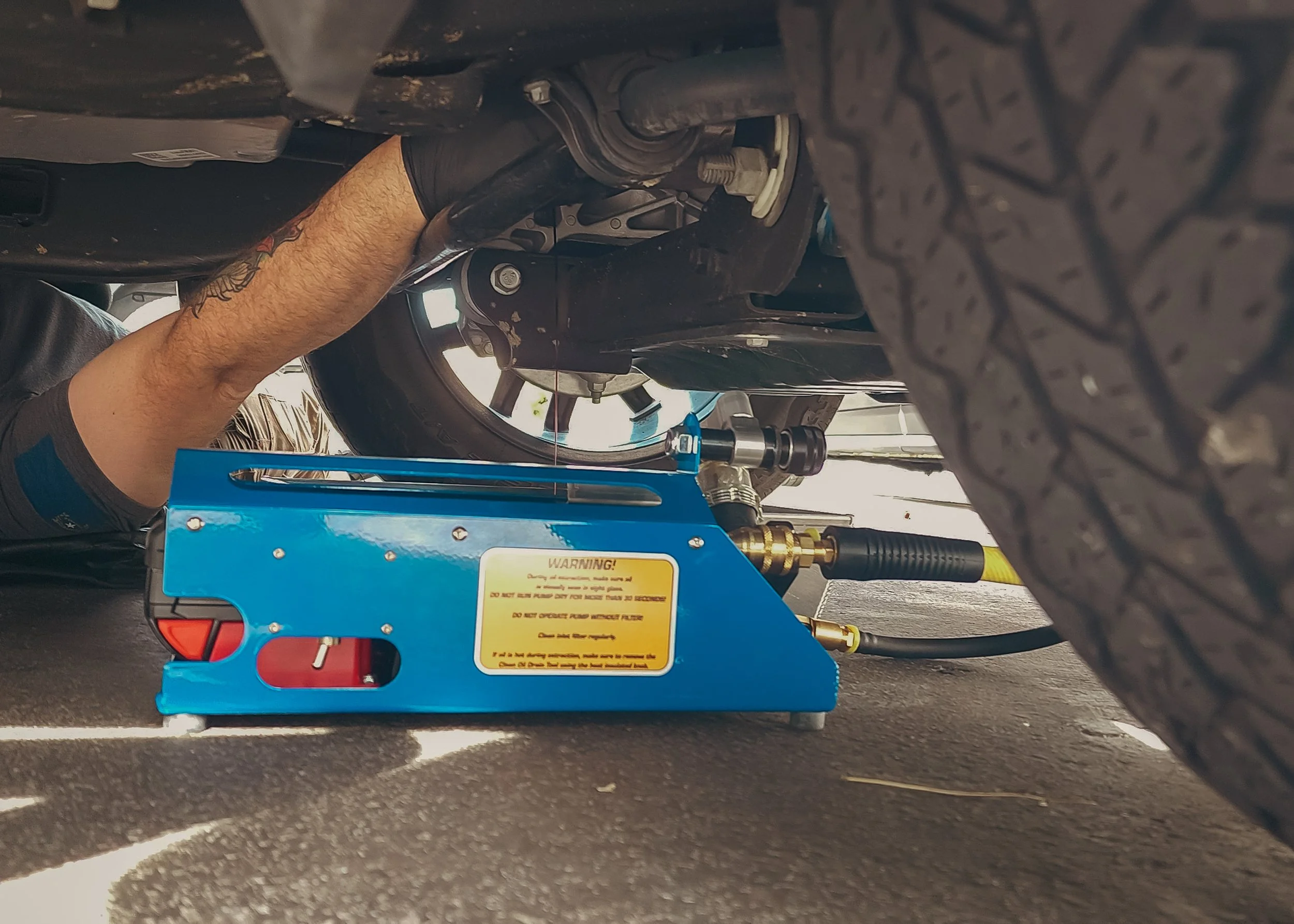 A person working underneath a vehicle using a blue hydraulic lift to lift the car, with tools and hoses connected to the car's underside.