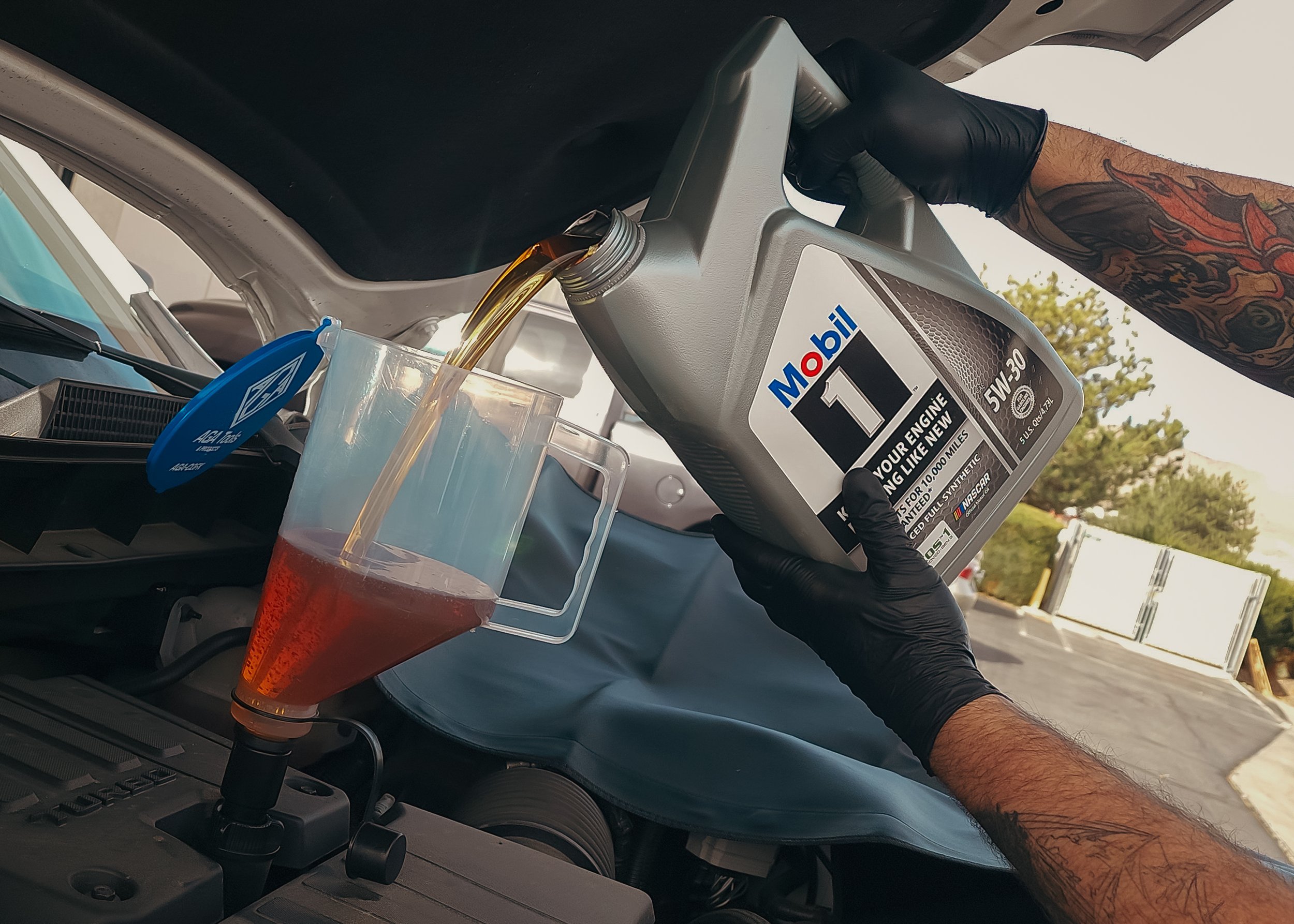 A person in black gloves pouring used motor oil from a plastic container into a clear plastic cup to recycle or dispose of properly, inside a vehicle with the hood open.