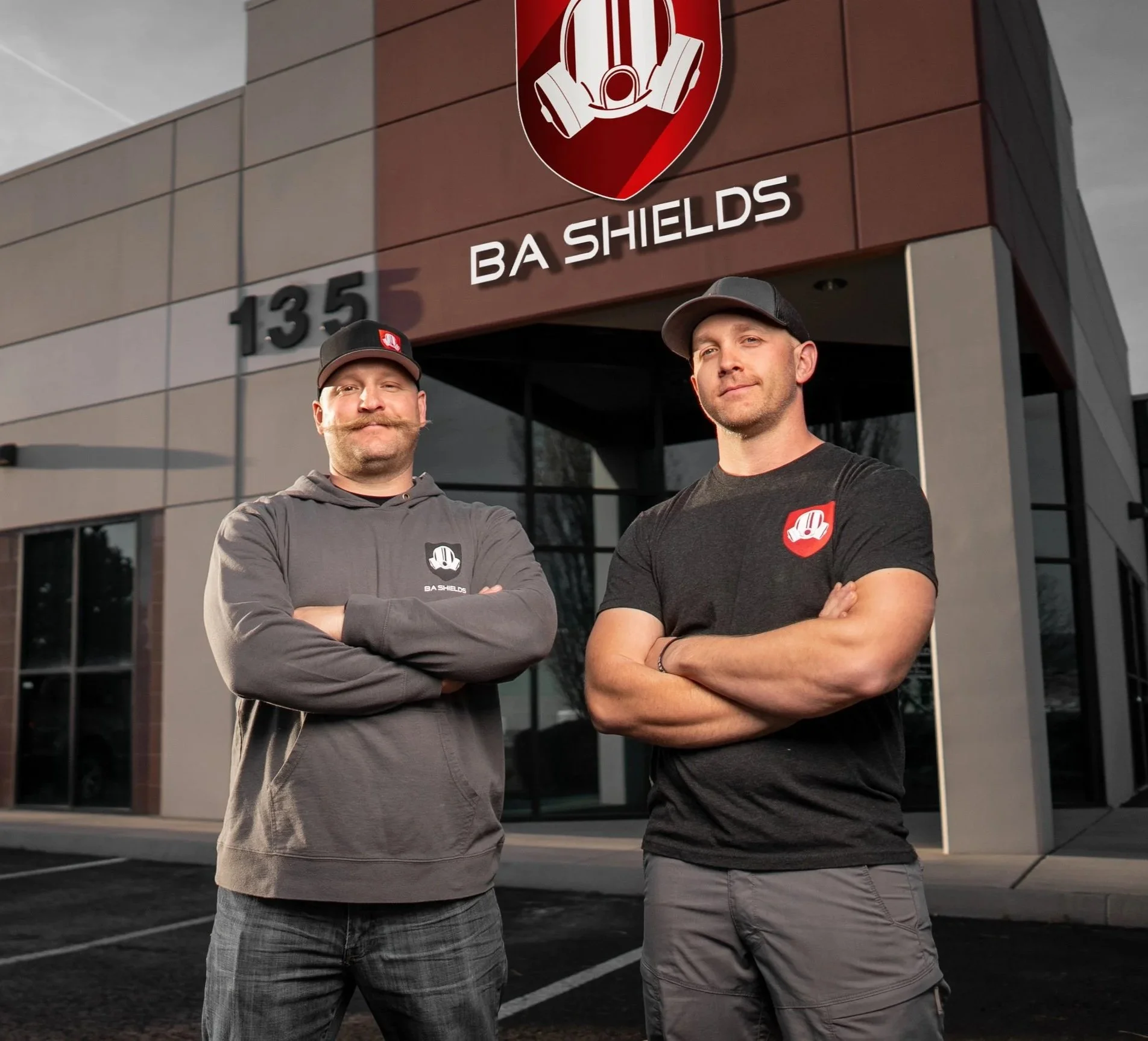 Two men standing outside a building with a sign that reads 'BA Shields,' both wearing casual clothing with the BA Shields logo, arms crossed, in front of the business's entrance.