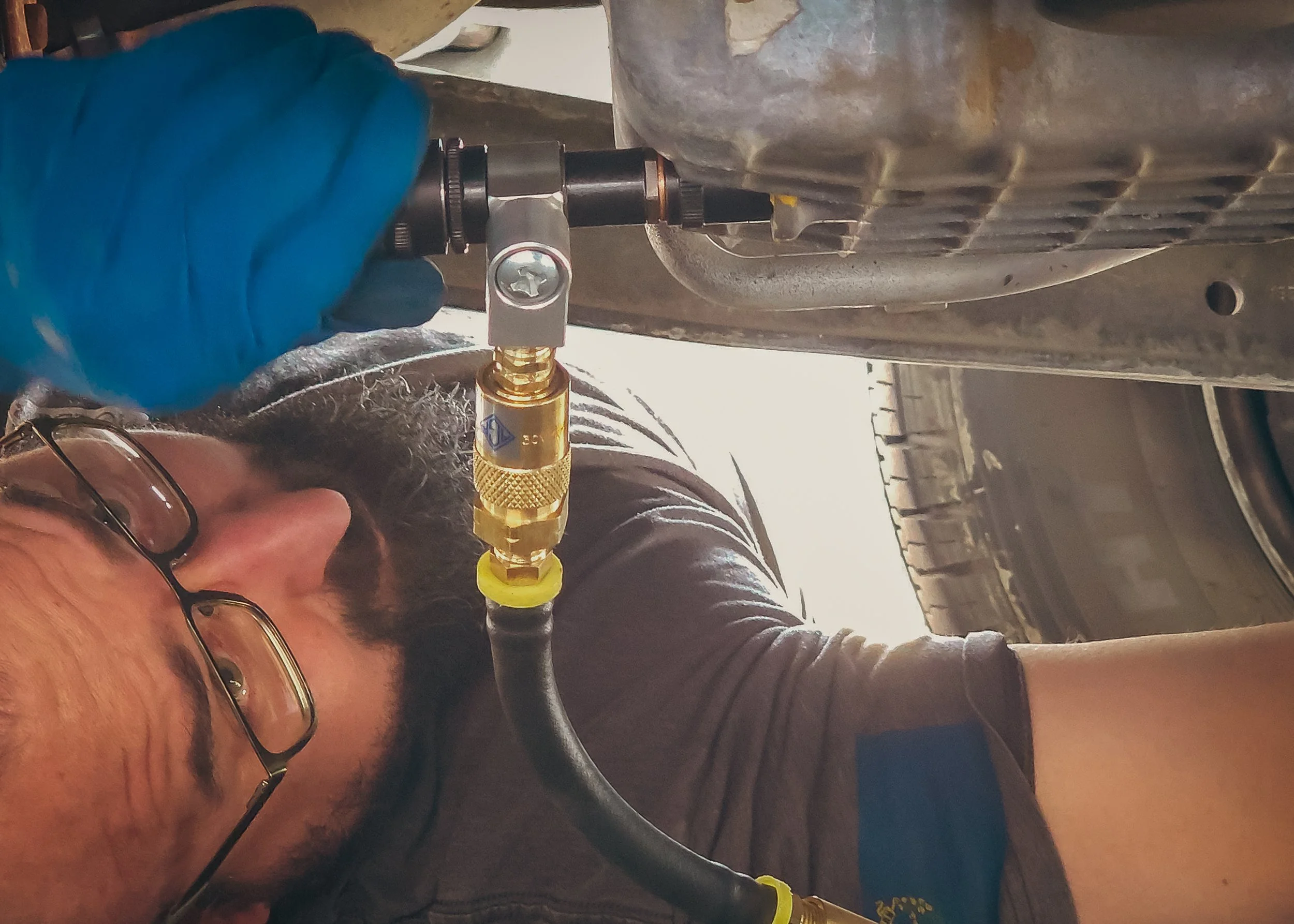 A man with glasses and a beard working underneath a vehicle, using a tool connected to the vehicle's oil pan.