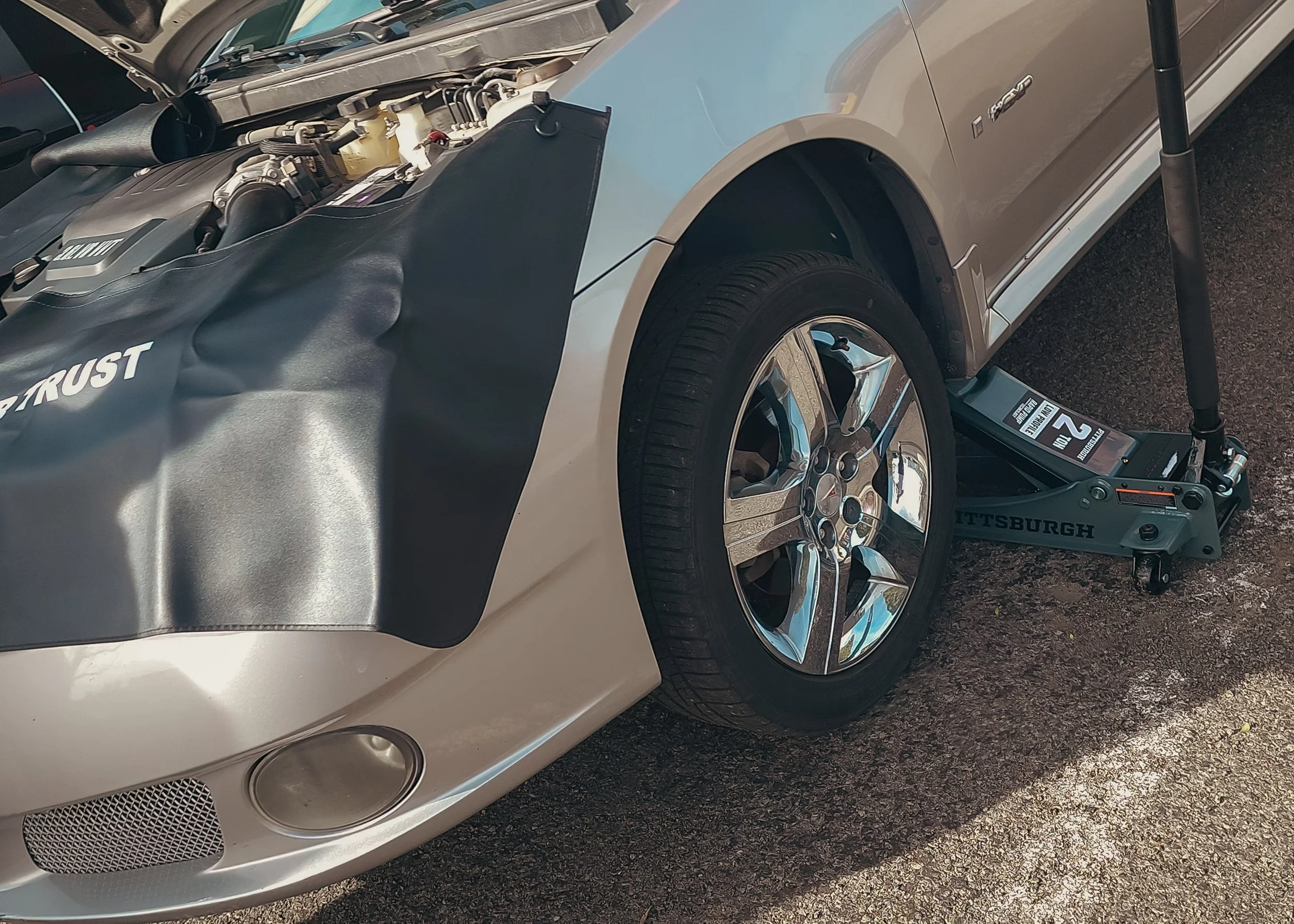 A silver car on a wheel lift. The hood cover with the word 'TRUST' partially visible is lifted, exposing the engine. The car is parked on asphalt.