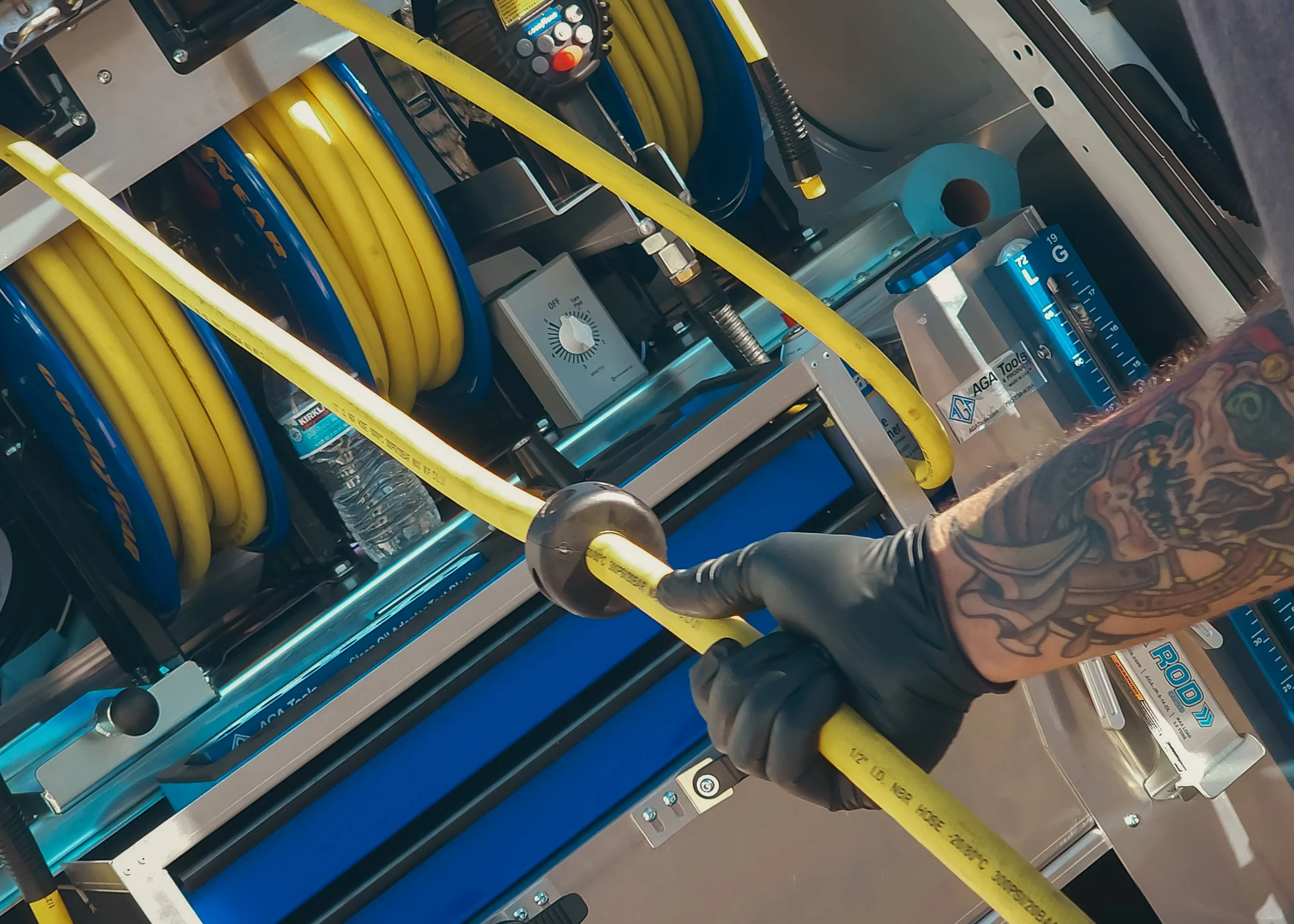 A person wearing a black glove is handling a yellow hose connected to equipment with blue and yellow hoses, on a metal control panel with a dial, surrounded by water bottles and tools in an industrial setting.