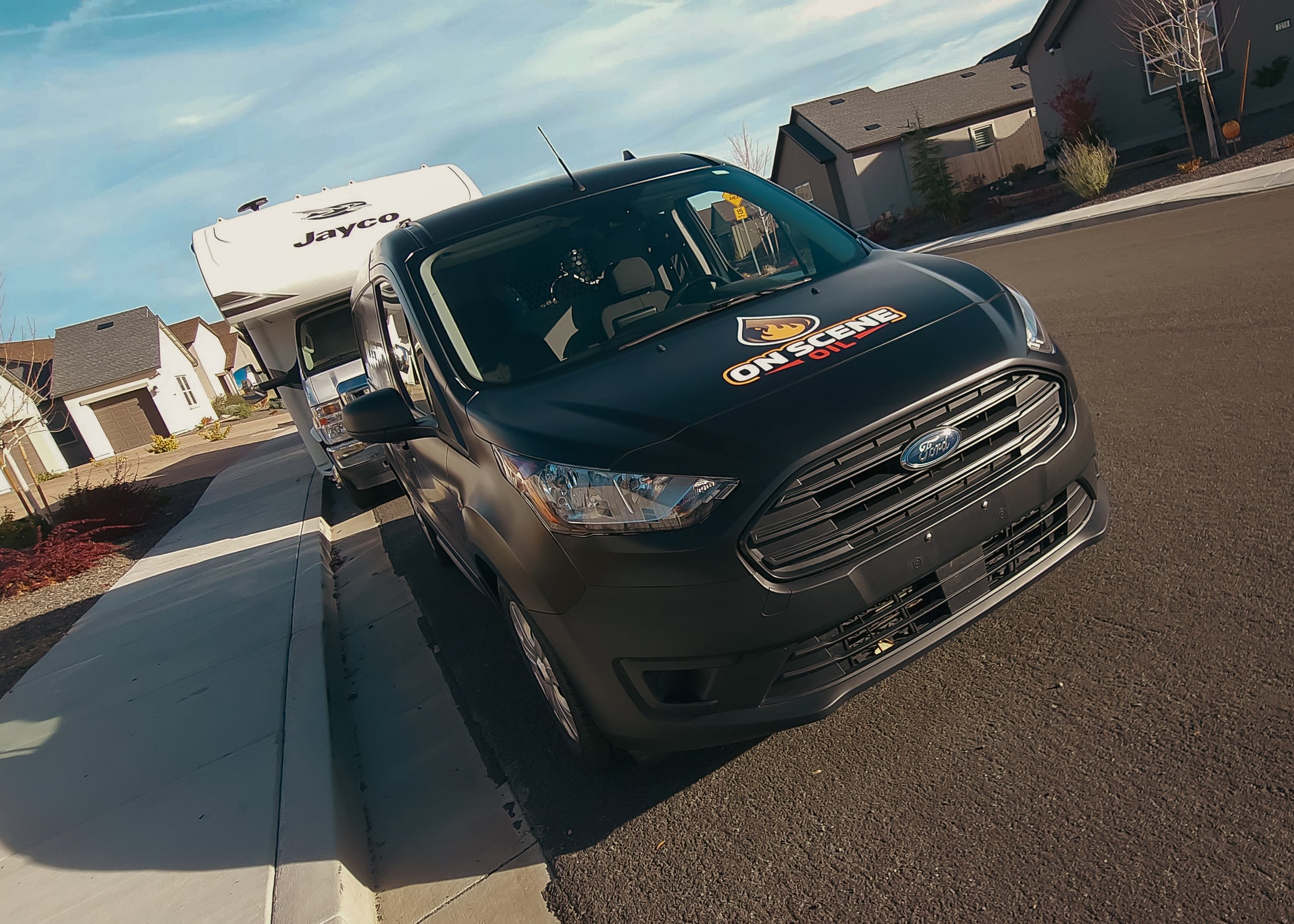 A black Ford transit van with a decal on the hood that reads 'ON SCENE OUT' and a man inside the vehicle, parked on a suburban street with houses and trees in the background.