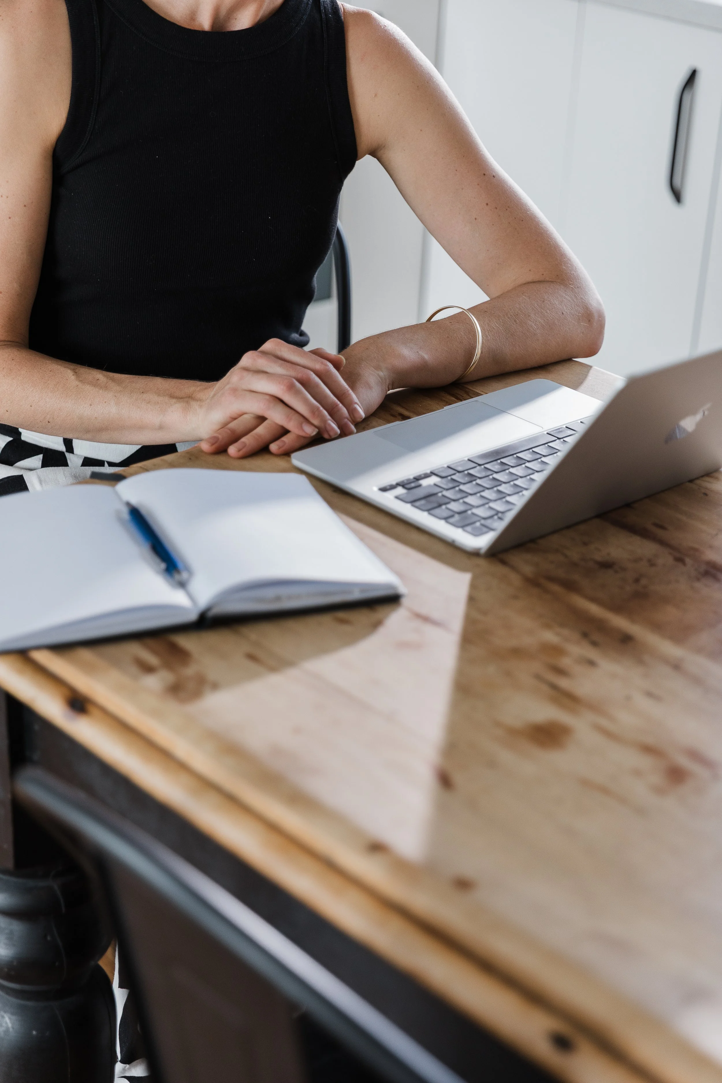 A woman sitting at a wooden table, working on a silver laptop, with an open notebook and pen nearby.