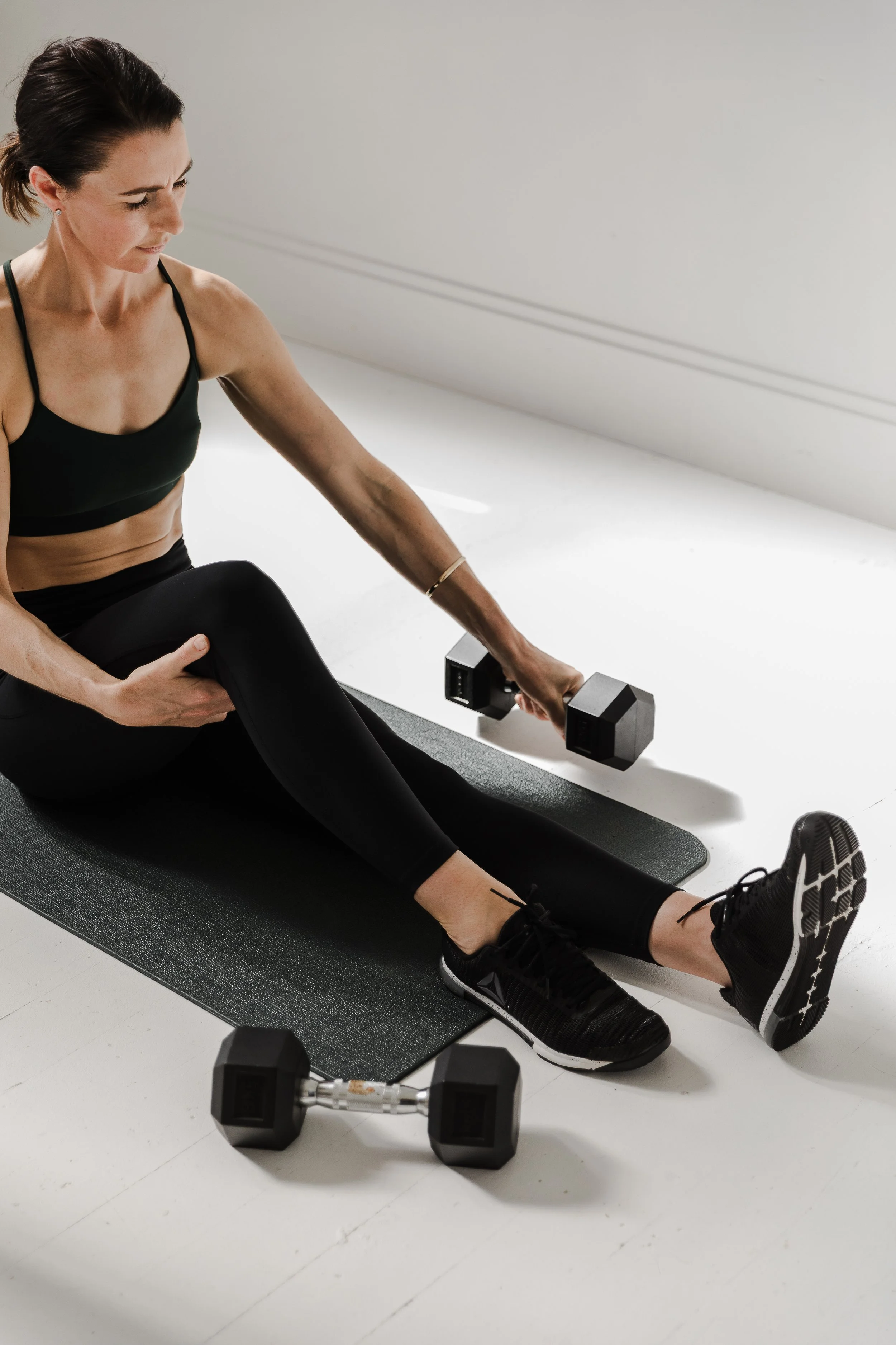 A woman in workout clothes sitting on a yoga mat, holding a dumbbell and stretching her leg. Dumbbells are on the floor nearby.