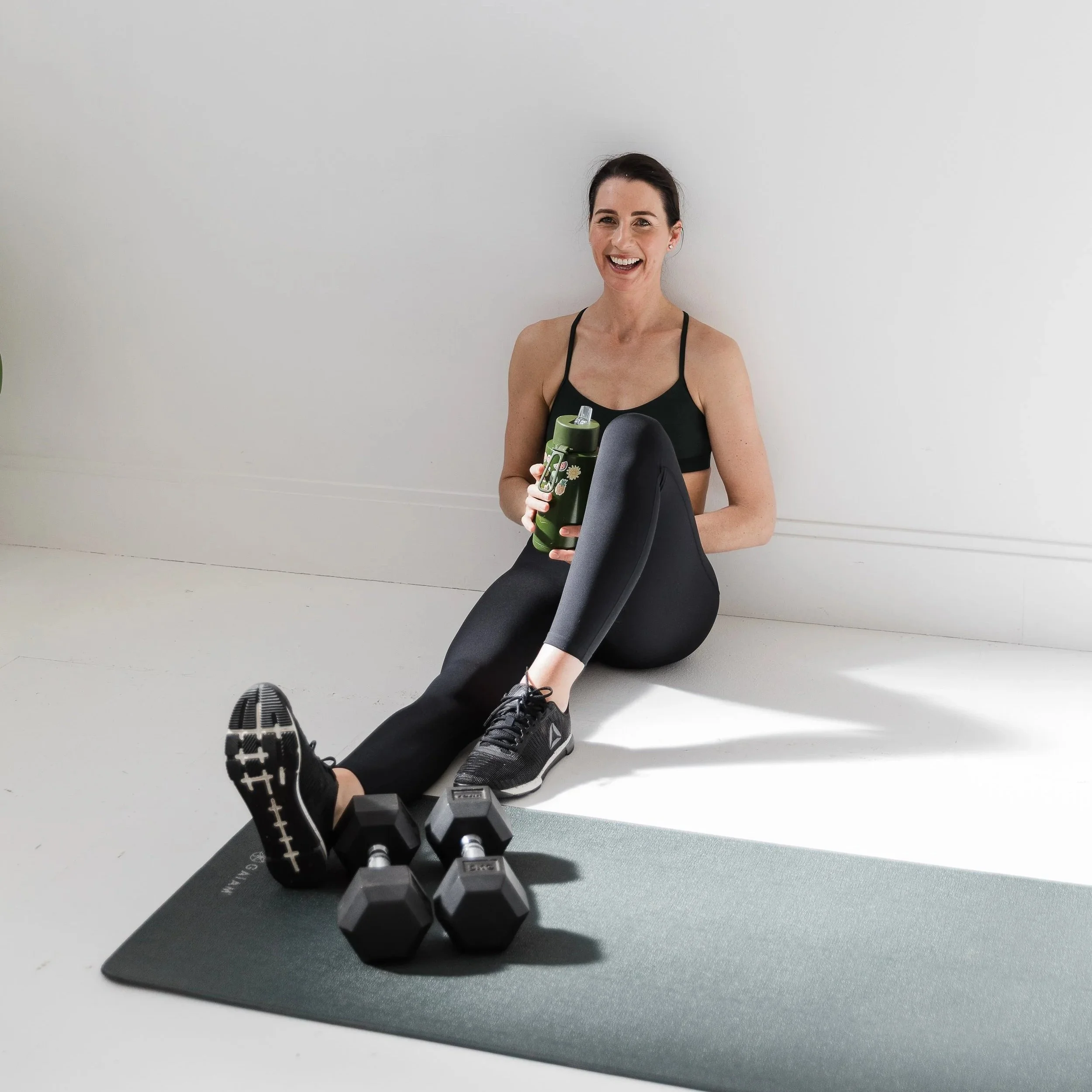A woman in workout attire sitting on a white floor, holding a green water bottle, with dumbbells and a workout mat in front of her, smiling at the camera.