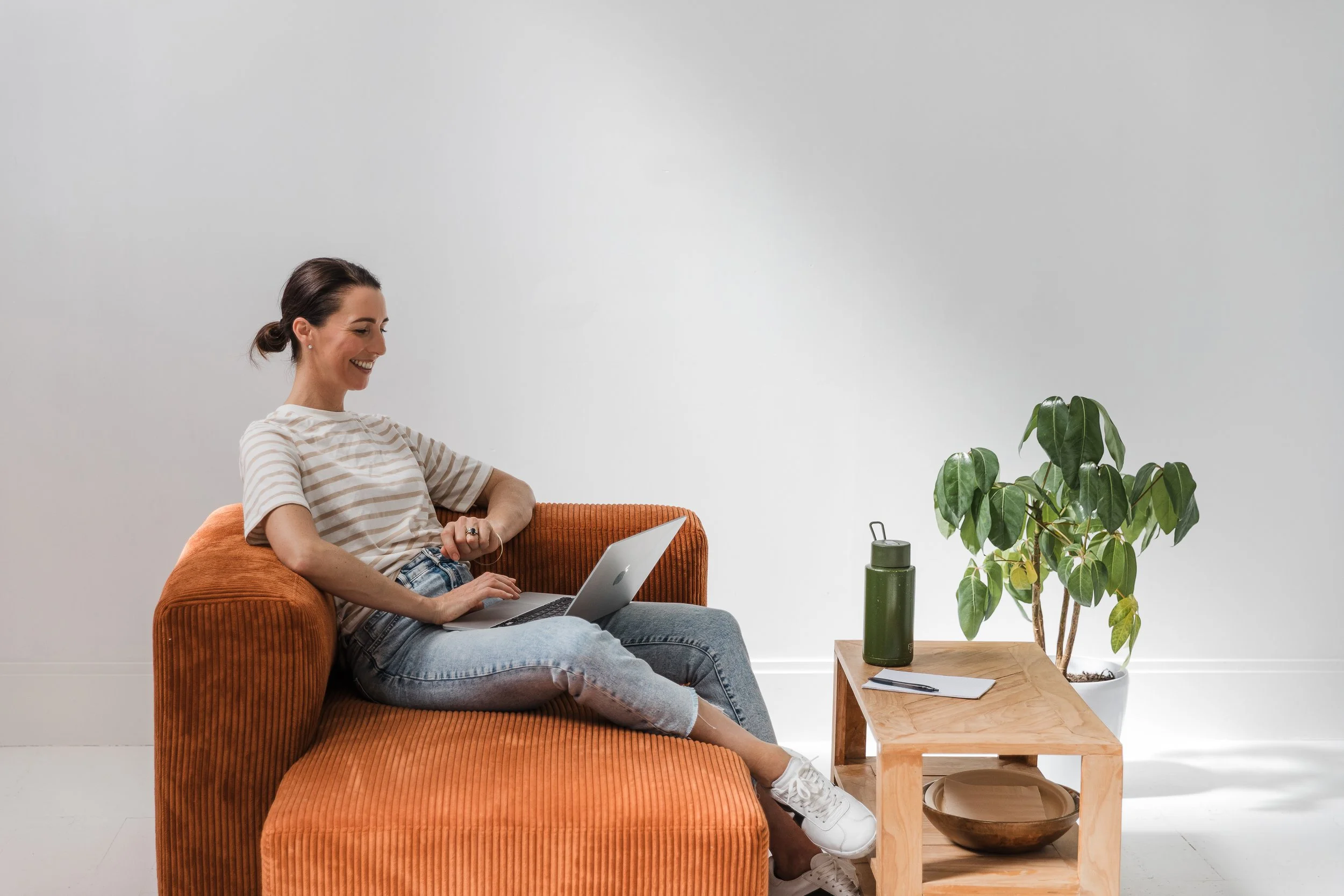 A woman sitting on an orange couch with a laptop, smiling, in a minimalist room with a wooden table and a potted plant.