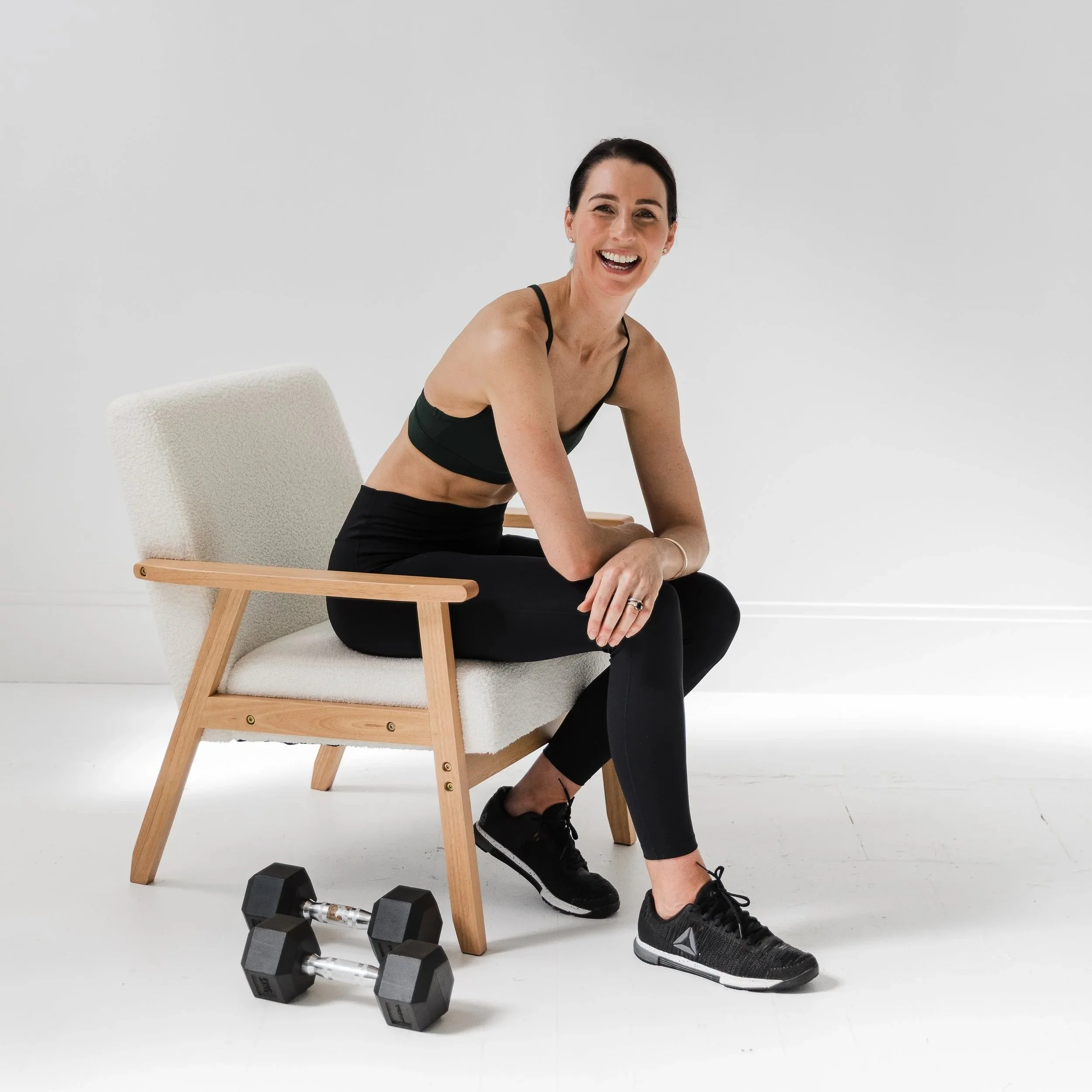 Woman dressed in workout attire sitting on a chair with dumbbells on the floor nearby, smiling at the camera.