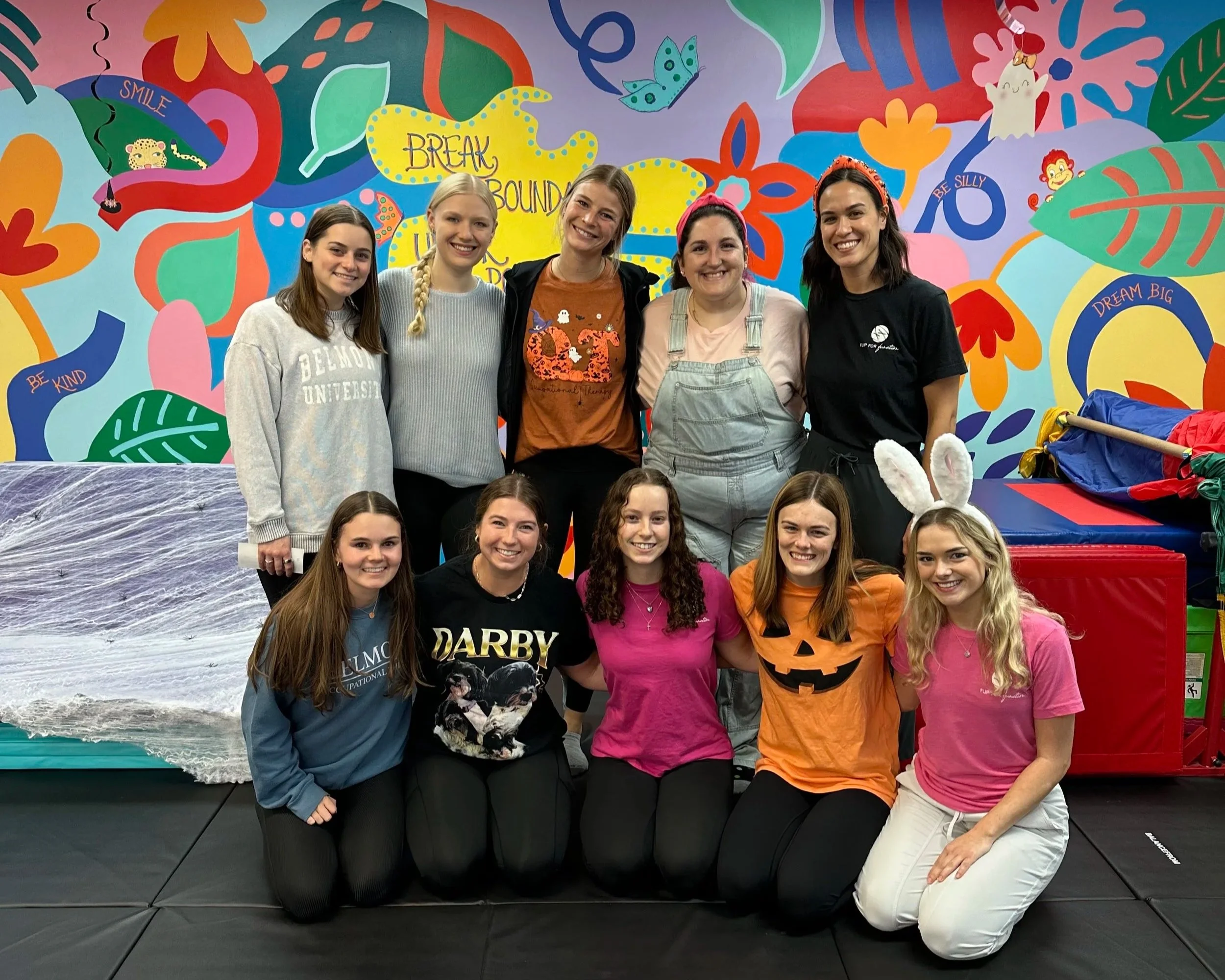 Group of young women posing together indoors, some with bunny ears, in front of a colorful mural with positive messages and playful designs.