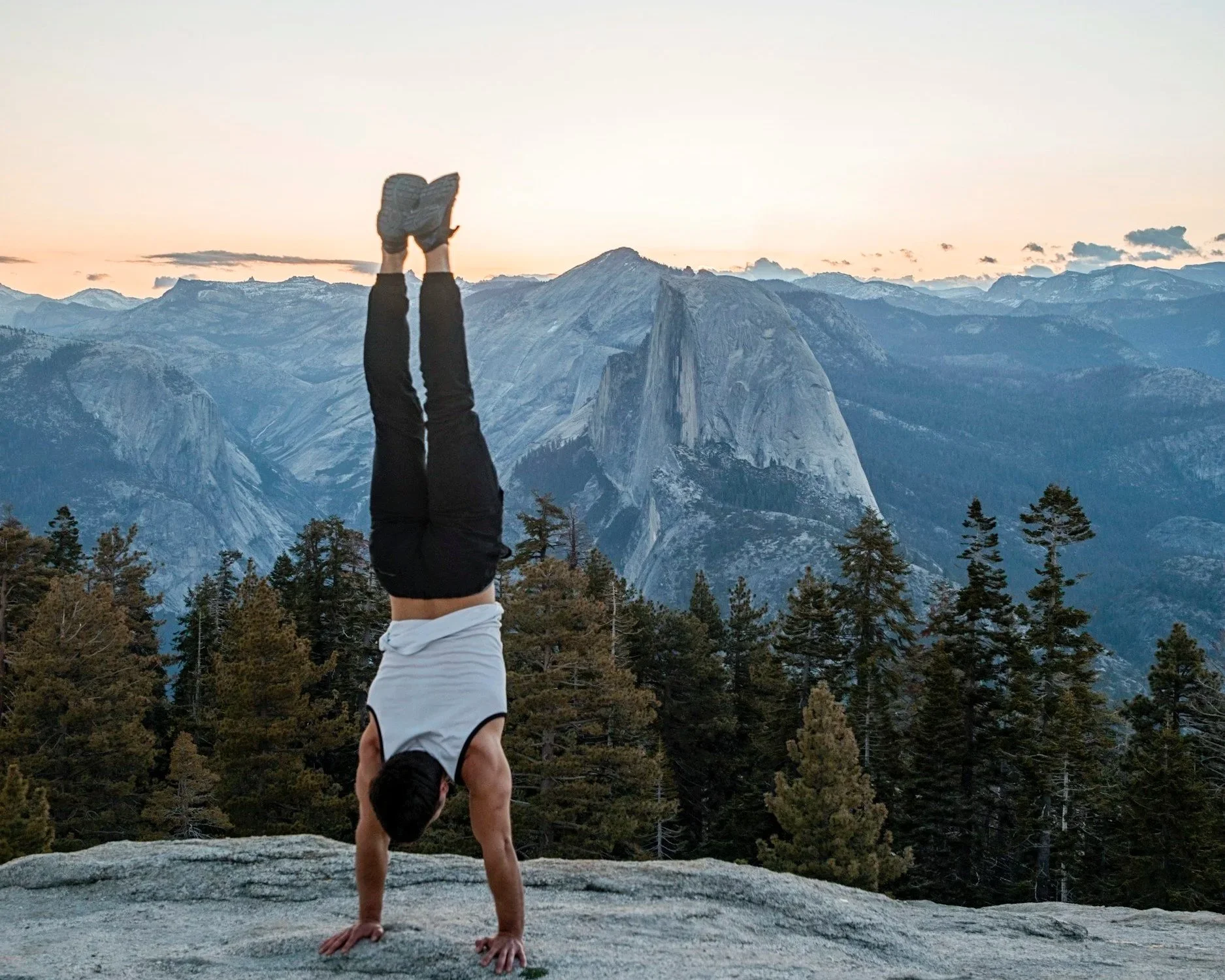 Person performing a handstand on a large rock with a mountain range and trees in the background during sunset.