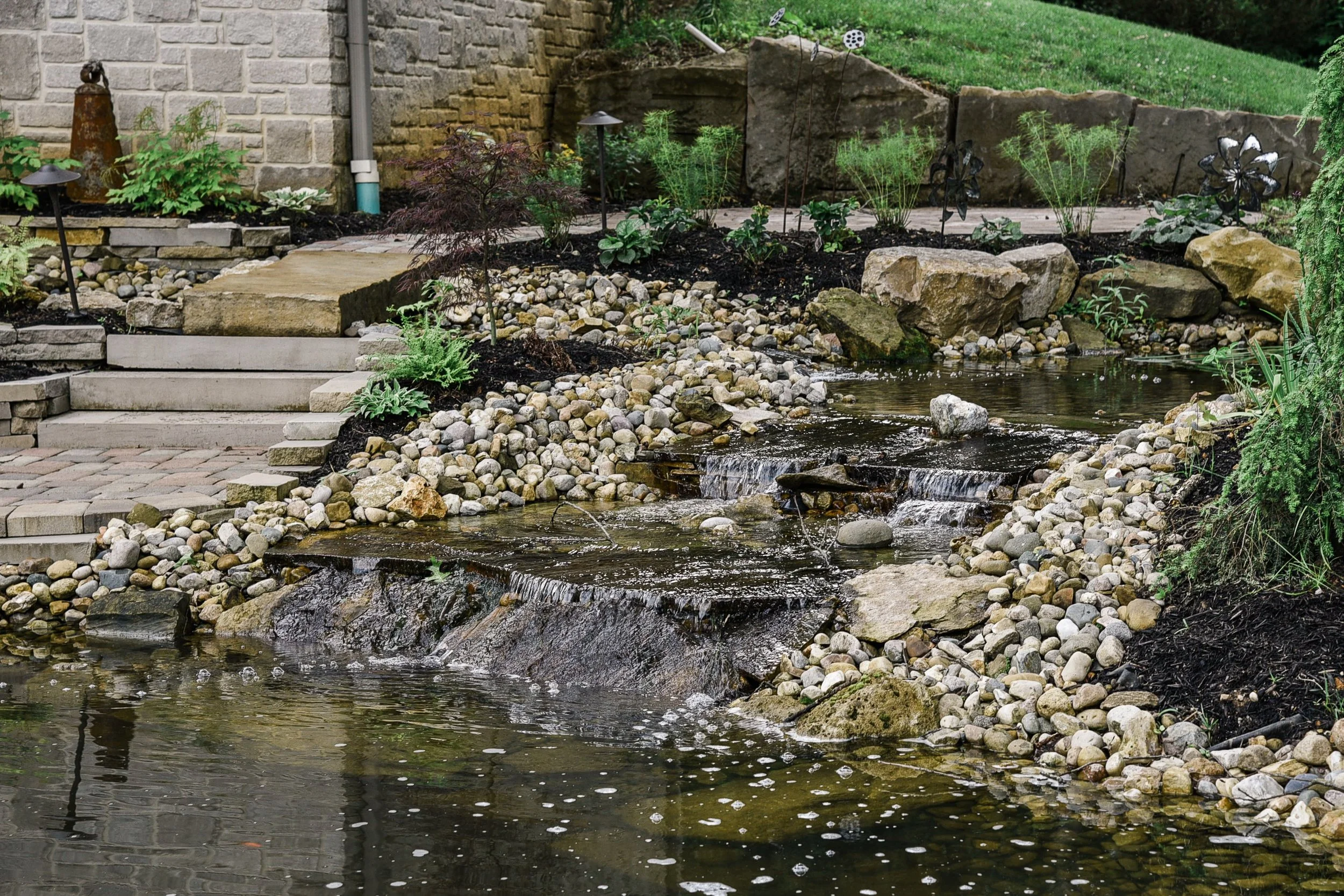 A backyard landscape with a small pond, rocks, plants, and garden lights.