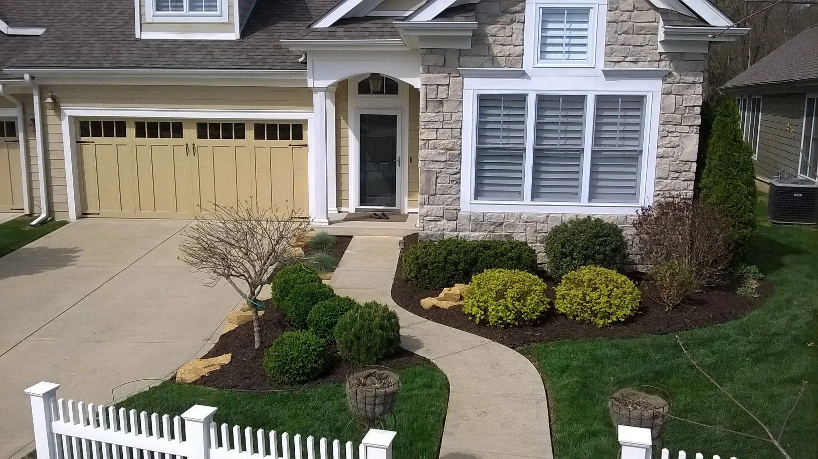 Front yard of a house with a landscaped garden, concrete walkway, small trees, bushes, and mulch beds, with a beige garage door and front entrance door.